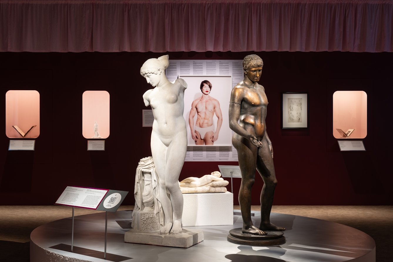 Photograph of a gallery view showing 2 prominent statues. A female figure in white stone on the left and a bronze male figure to the right. Between them, hung on the far gallery wall is a photographic self-portrait by the artist, Cassils.