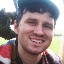 Colour photograph headshot of Andrew Kerr wearing a hat and plaid shirt and smiling.