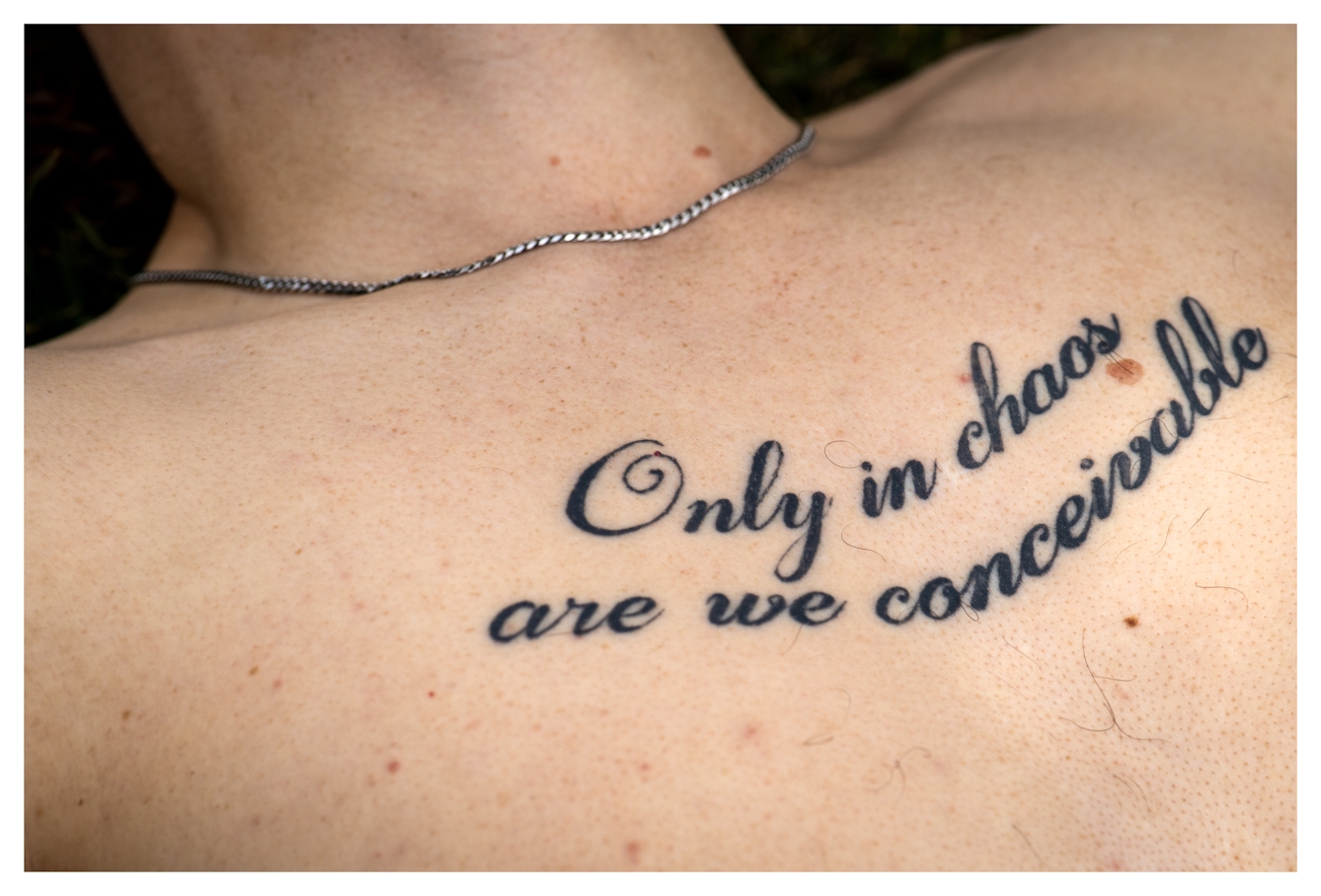 Photograph of the top section of a naked man's chest, from his sternum to his neck. Across his chest is a tattoo in ornate script with the words "Only in chaos are we conceivable". Around his neck is a simple silver chain necklace.