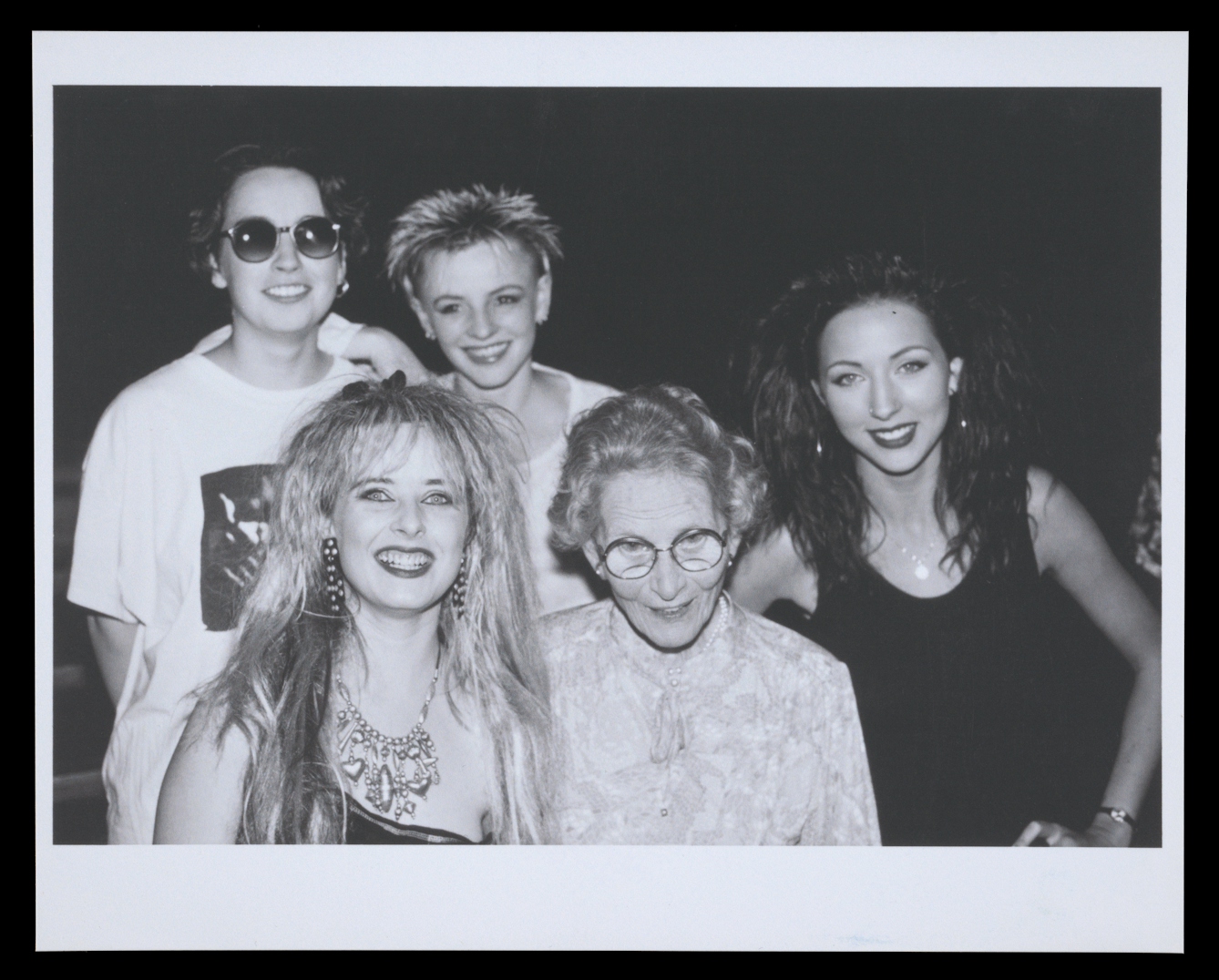 Photograph of a black and white archive photographic print from the 1980s against a black background. The print shows a group photograph of 4 young people surrounding an older woman who is wearing glasses. Everyone is looking to camera and smiling.