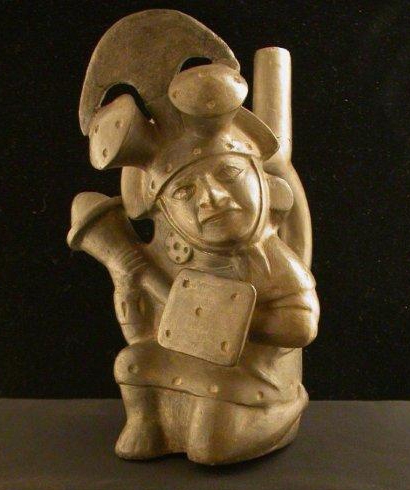 Photograph of a ceramic sculpture of a warrior, kneeling on left knee, bearing square shield on left arm and massive club in right hand. Wears elaborate helmet with crosswise crest, crescentic blade and two mushroom knobs.