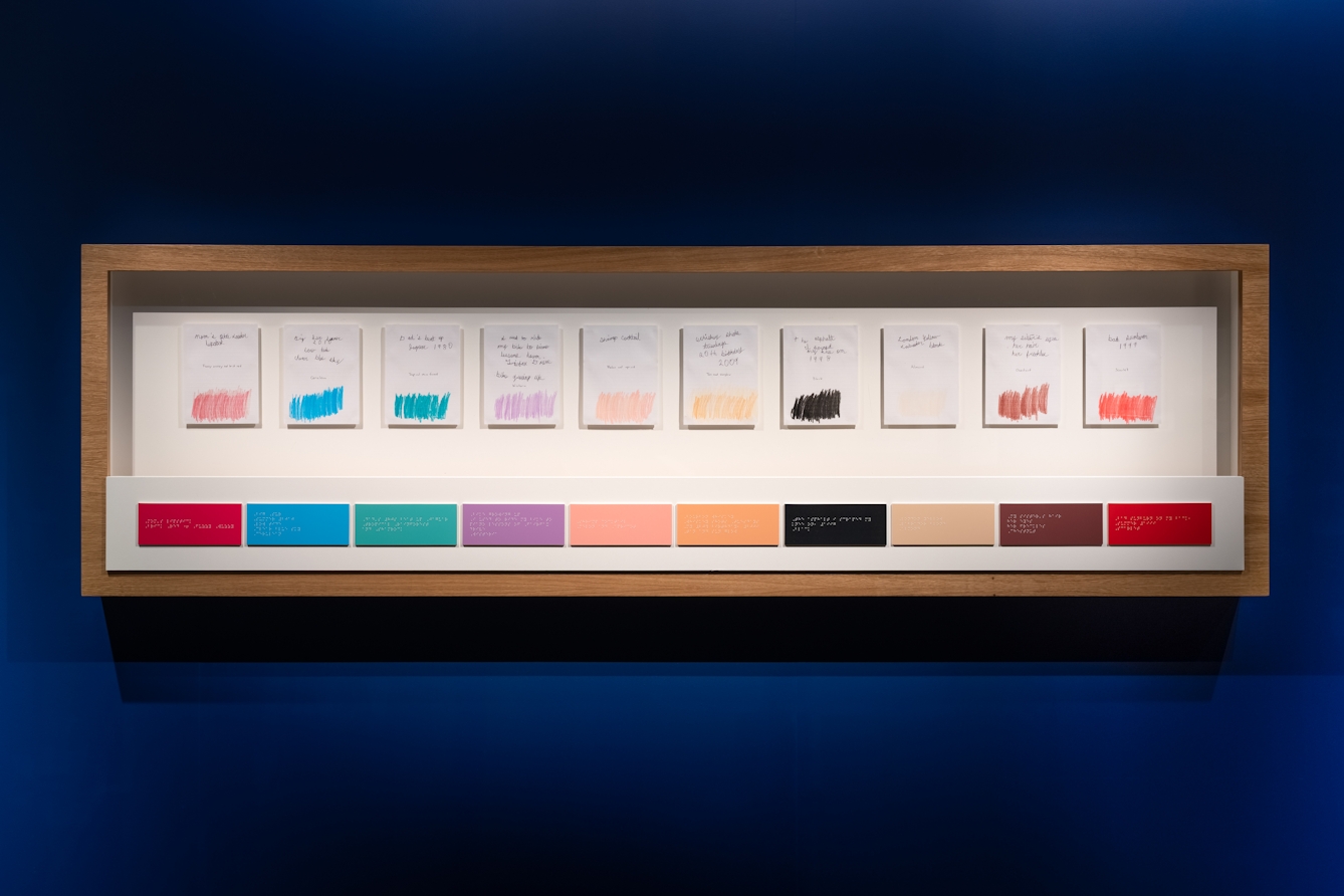 Photograph of a framed artwork on a blue gallery wall. The wooden frame contains a row of ten colour rectangular swatches ranging in hues including red, blue, purple, pink and black. Above each of these swatches is a sheet of paper with the same colour sketched out roughly by hand. Each is accompanied by some handwritten text.