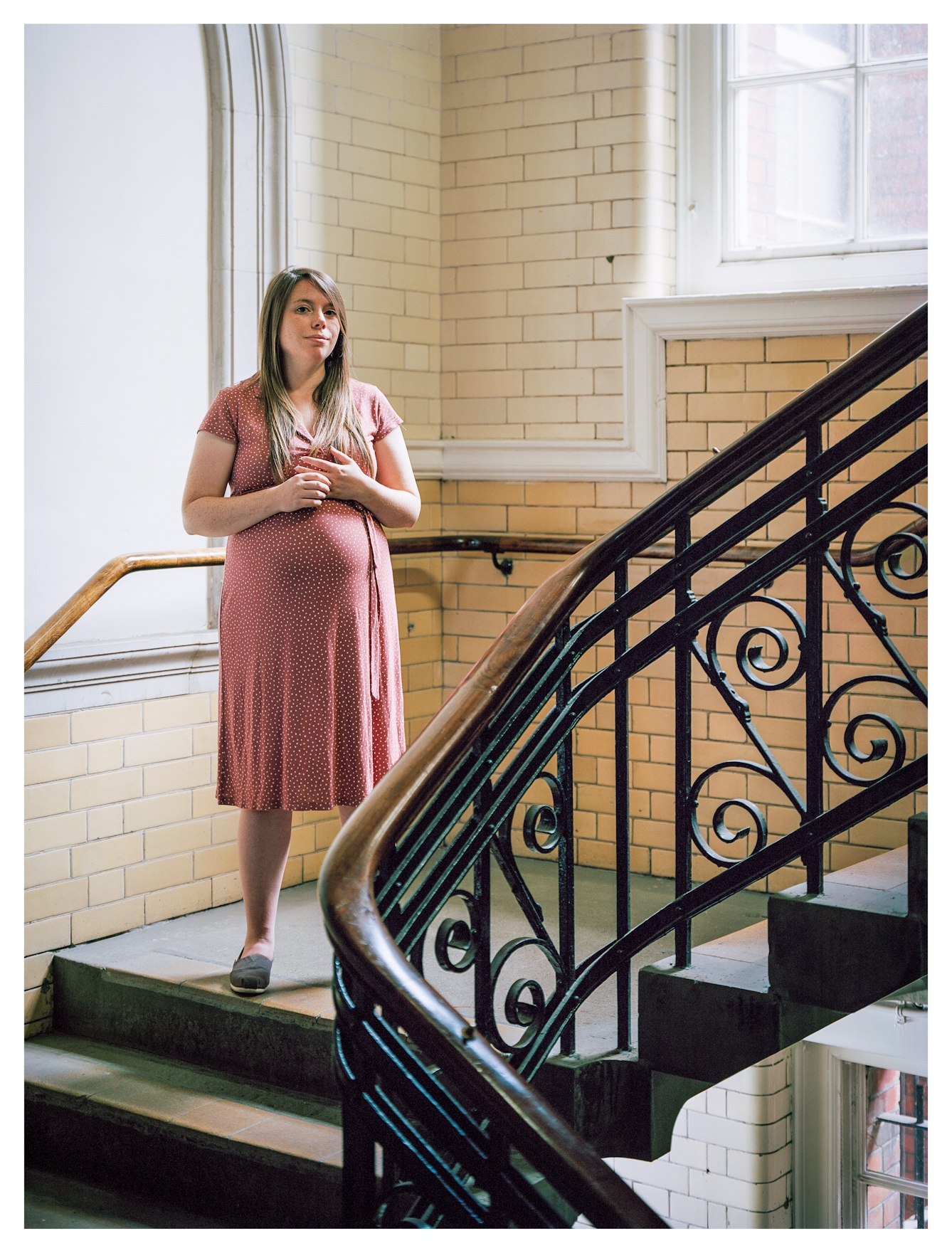 Photographic full length portrait of a woman standing looking to camera with her hands clasped in front of her chest. She is wearing a red dress. She is lit by a soft light from a large window to the right. Behind her are the glossy tiled walls of a Victoria period building and a large staircase with case iron balustrades made in intricate curls.