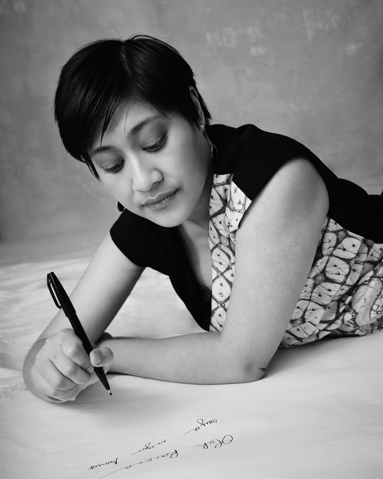 Picture in black and white of Okka writing in Indonesian on a paper floor with a pen. She’s wearing a patterned black and batik dress, with short hair and black and silver earrings. If you’re using this image, kindly credit photographer Derrick Kakembo.