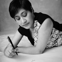 Picture in black and white of Okka writing in Indonesian on a paper floor with a pen. She’s wearing a patterned black and batik dress, with short hair and black and silver earrings. If you’re using this image, kindly credit photographer Derrick Kakembo.