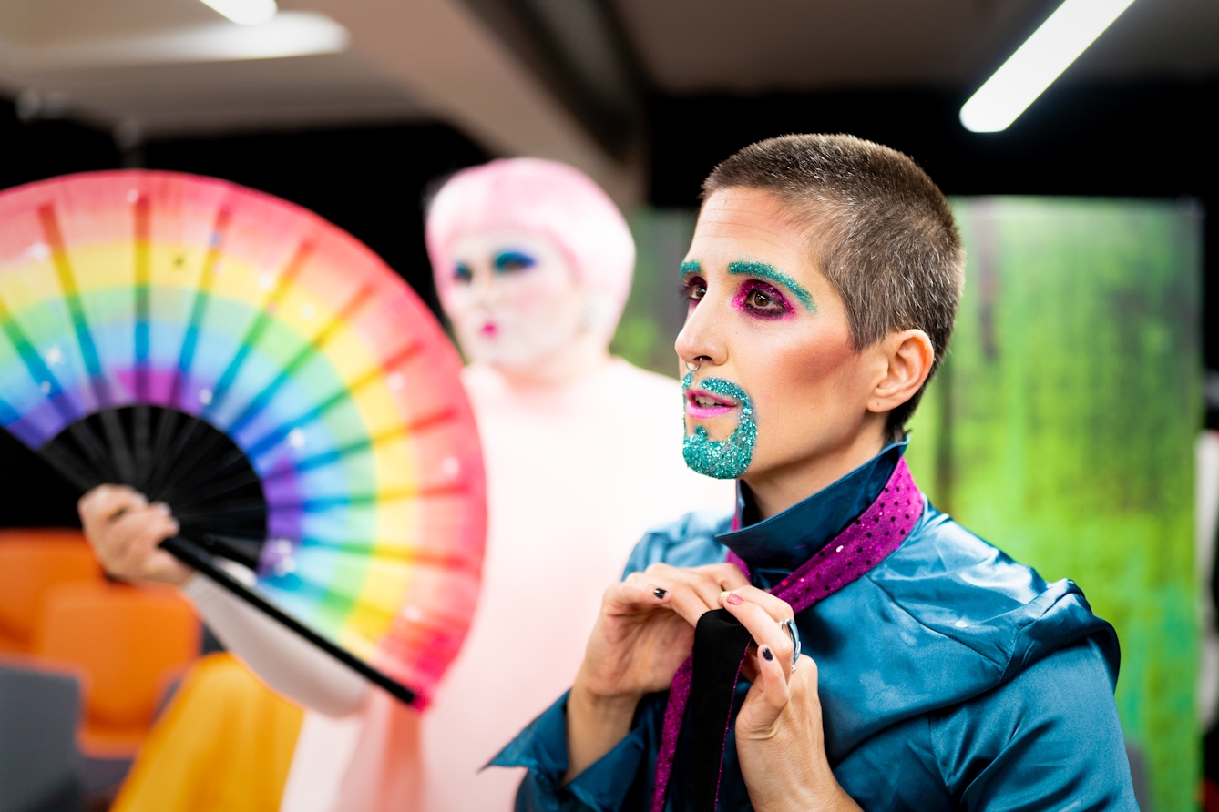 Photograph showing a performer backstage. The performer is dressed in a shiny blue shirt. They are in the process of tying their purple tie. Their mouth and eye brows are covered in blue glitter make-up. In the background is another performer holding a large rainbow coloured hand fan.