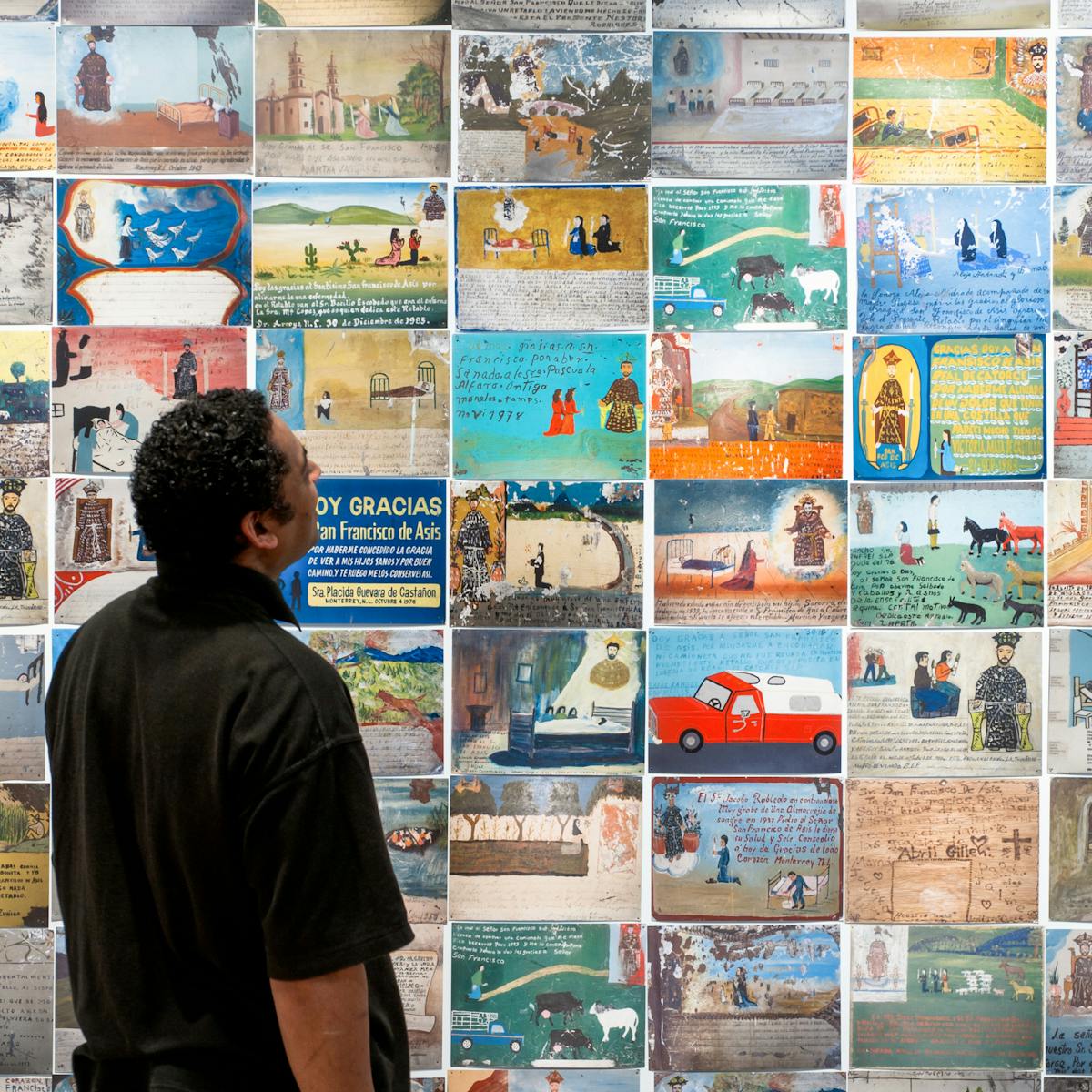 Photograph of a man looking at a wall of colourful Mexican votives, part of the exhibition Infinitas Gracias: Mexican miracle paintings at Wellcome Collection.