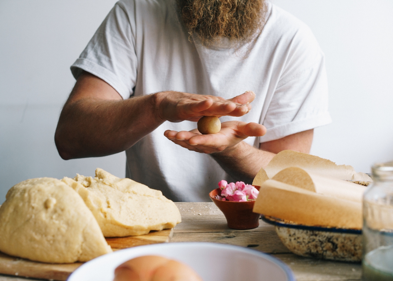 Photograph of a man sat at a wooden kitchen table. Only his torso can be seen along with part of his long beard. He is wearing a white t-shirt. On the tabletop is a dough mixture, a small bowl of flower heads and baking trays lined with greaseproof paper. The man is in the process of rolling the dough into small ball shapes with his hands.