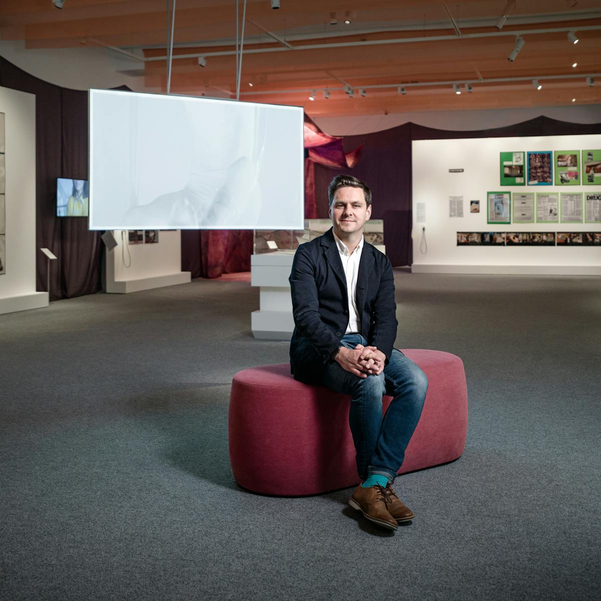 Photograph of Misbehaving Bodies curator George Vasey sitting in the exhibition space.  Behind him are the works of Jo Spence; in the form of photographs and tear sheets hung on white panels, and Oreet Ashery; in the form of a projection of a white figure moving against a white screen.