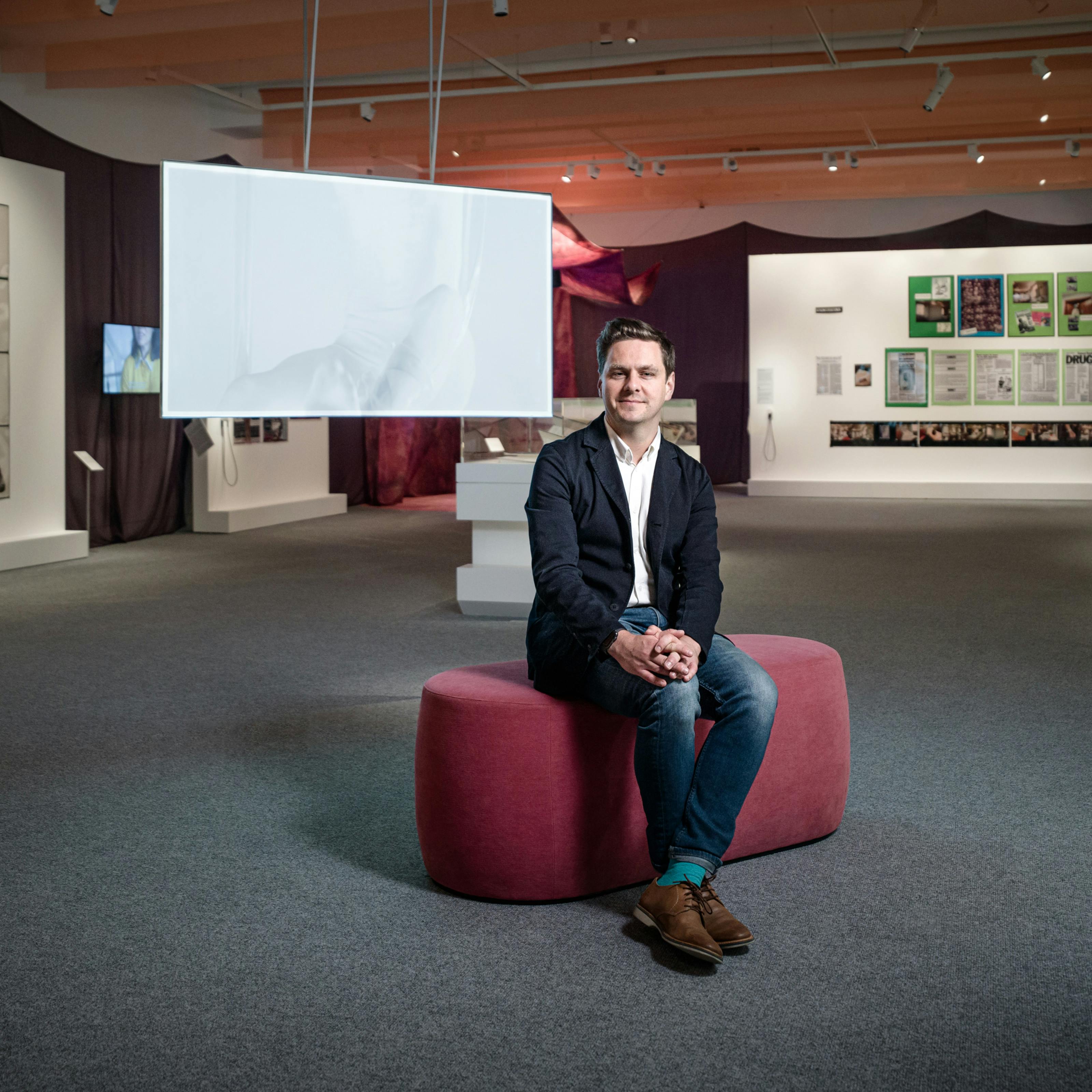Photograph of Misbehaving Bodies curator George Vasey sitting in the exhibition space. Behind him are the works of Jo Spence; in the form of photographs and tear sheets hung on white panels, and Oreet Ashery; in the form of a projection of a white figure moving against a white screen.