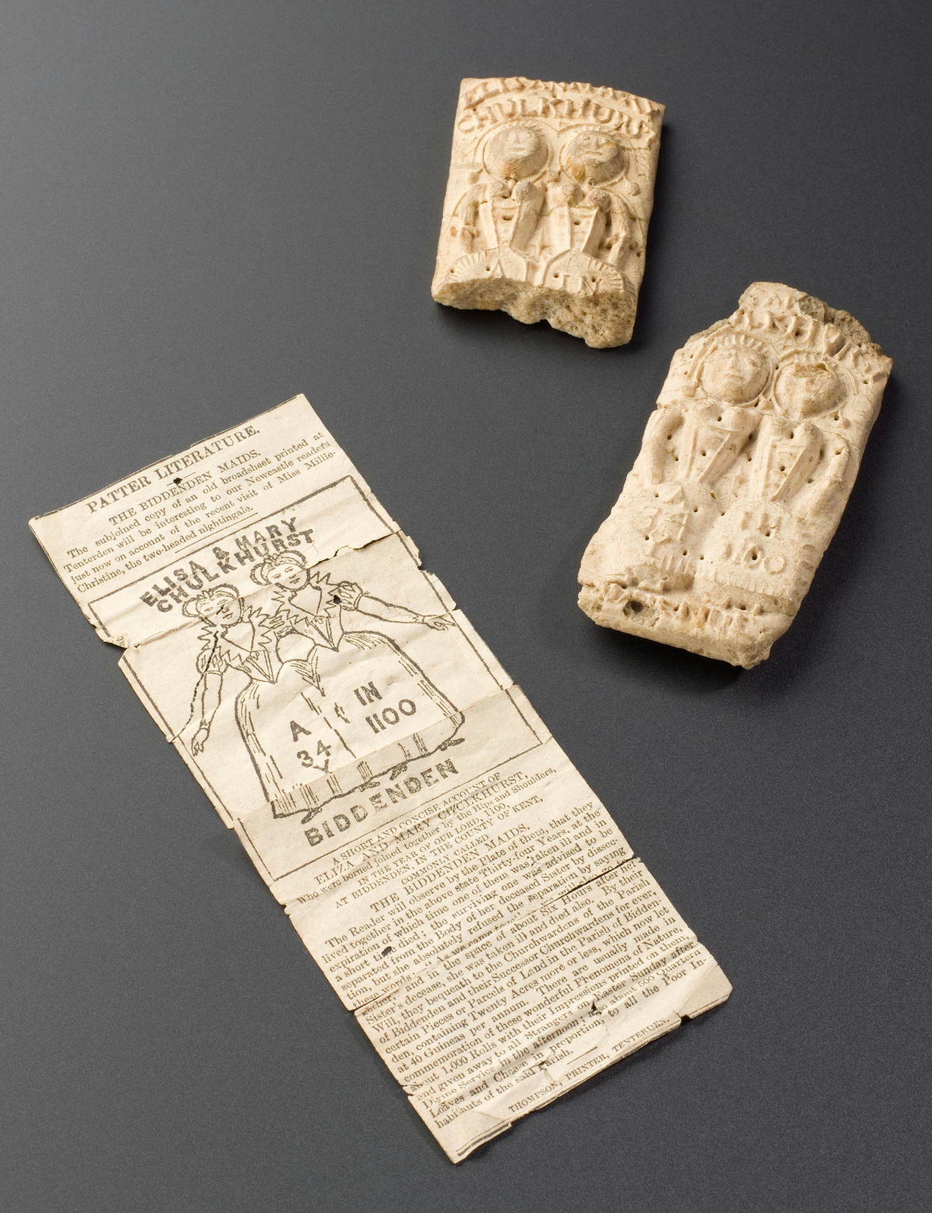 Photograph showing a pamphlet and two biscuits resting on a grey background. 

The pamphlet has an image of a pair of conjoined twins and text which reads 'Elisa and Mary Chulkhurst. Biddenden.' The rest of the text is illegible. The biscuits have the same image of the twins embossed onto them, one of the biscuits has been broken.