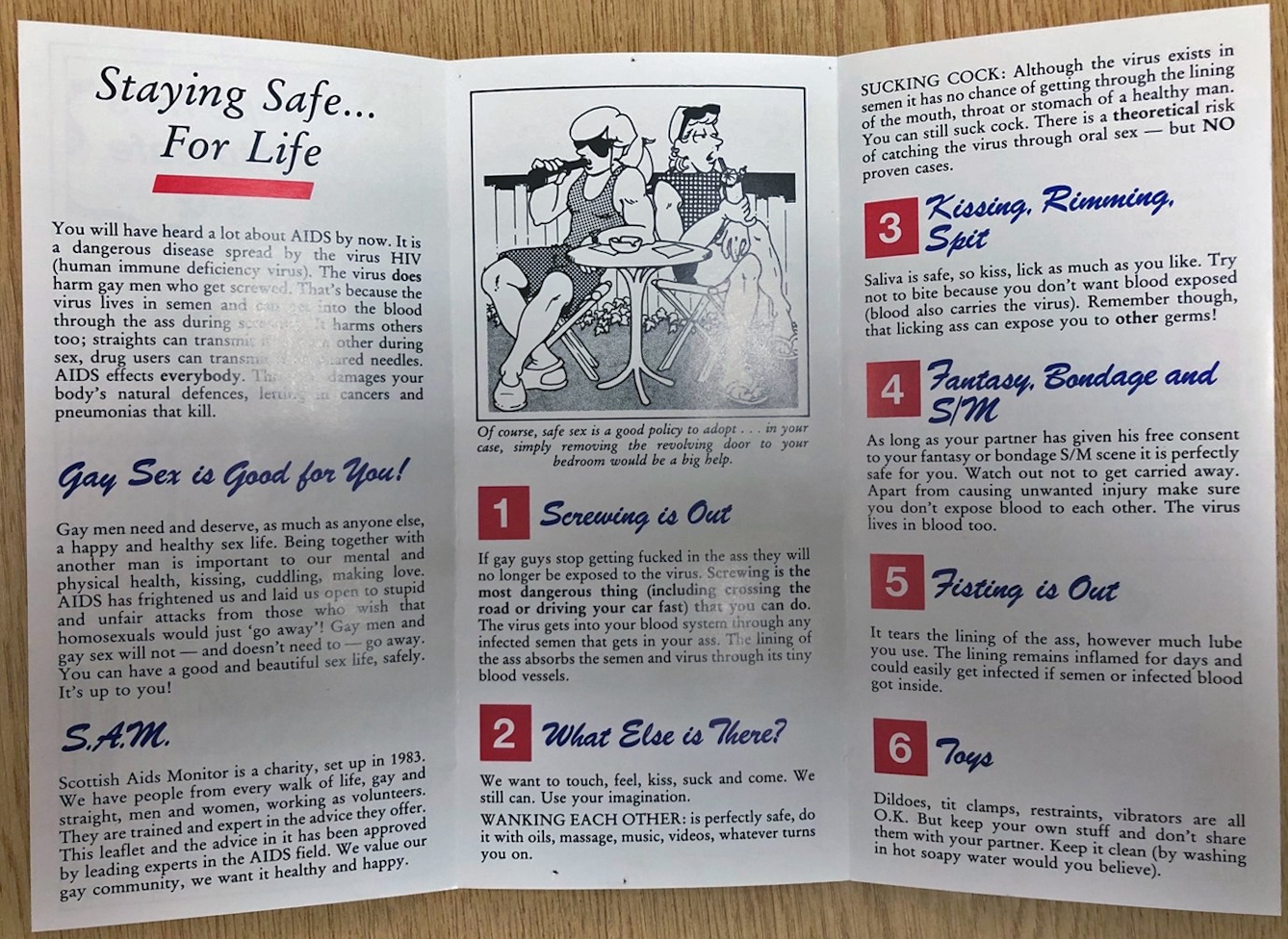 Photograph of the inside of a pamphlet folded in three. The title is "Staying Safe... For Life". In the middle section, there is an image of two women chatting at an outside table, with the caption "Of course, safe sex is a good policy to adopt... in your case, simply removing the revolving door to your bedroom would be a big help." There is information in the pamphlet about the Scottish Aids Monitor and various sexual practices such as fisting, rimming, bondage and toys.