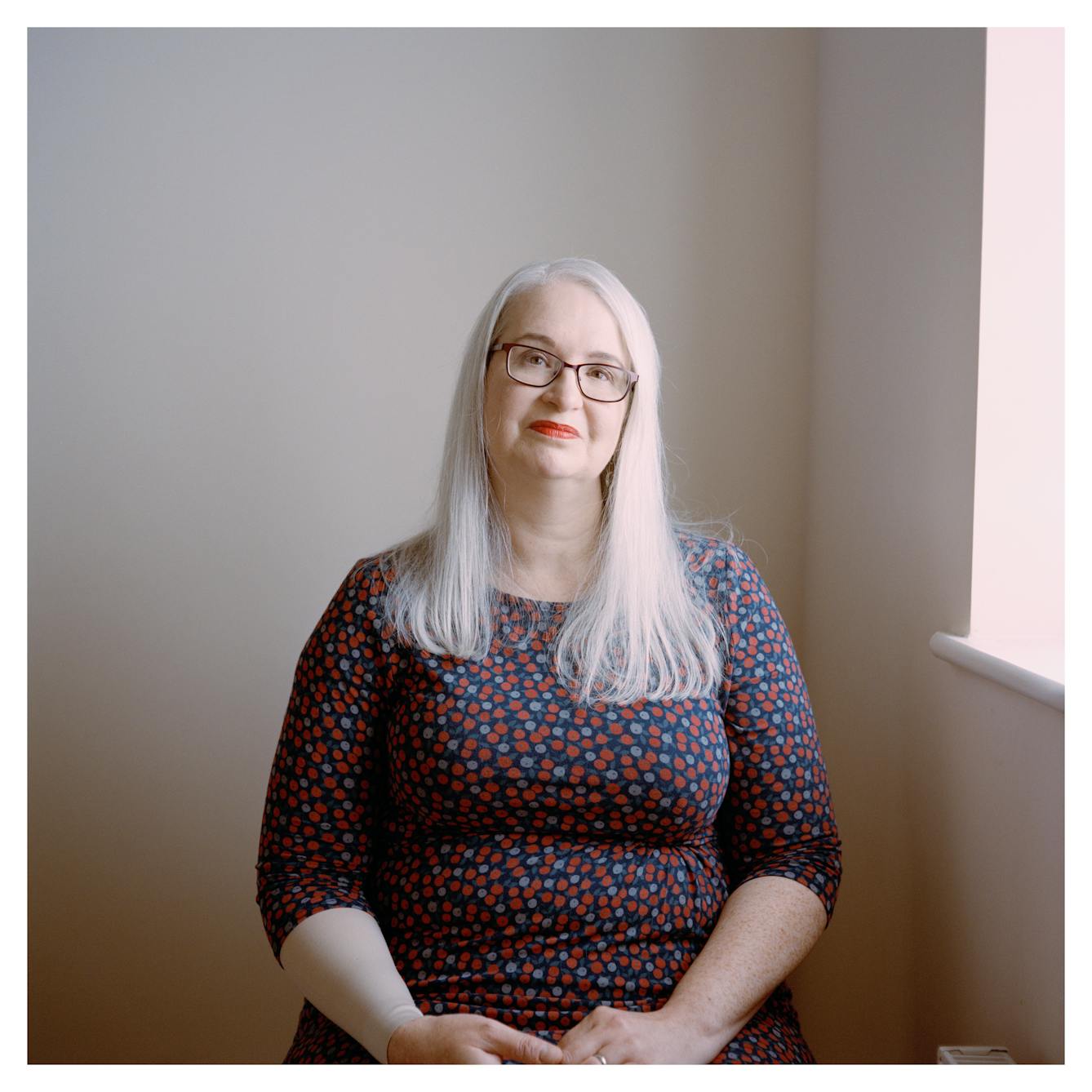Portrait photograph of Louise, a woman with long grey hair. She is sitting down and looking directly at the camera with a neutral expression. 

She is wearing black rectangular glasses, red lipstick and a blue and red floral patterned dress. Her hands are clasped on her lap. There is a white wall behind her, and a window ledge. 