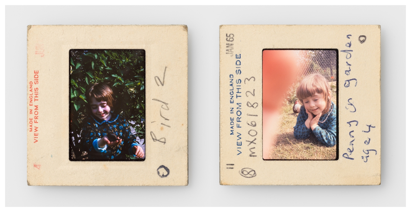 Photograph of two colour 35mm transparencies side by side, each mounted in a cardboard side holder, resting on a white background. The transparency on the left shows a young girl in a dark blue dress standing in green foliage, holding a small bird in her right hand. The slide mount has the words 'made in England. View from this side' printed on it. A hand written note on the mount reads, "Bird 2". The transparency on the right shows the same young girl in a blue dress lying on her front on a lawn. The slide mount has the words 'made in England. View from this side' printed on it. A hand written note on the mount reads, "Penny in garden aged 4".