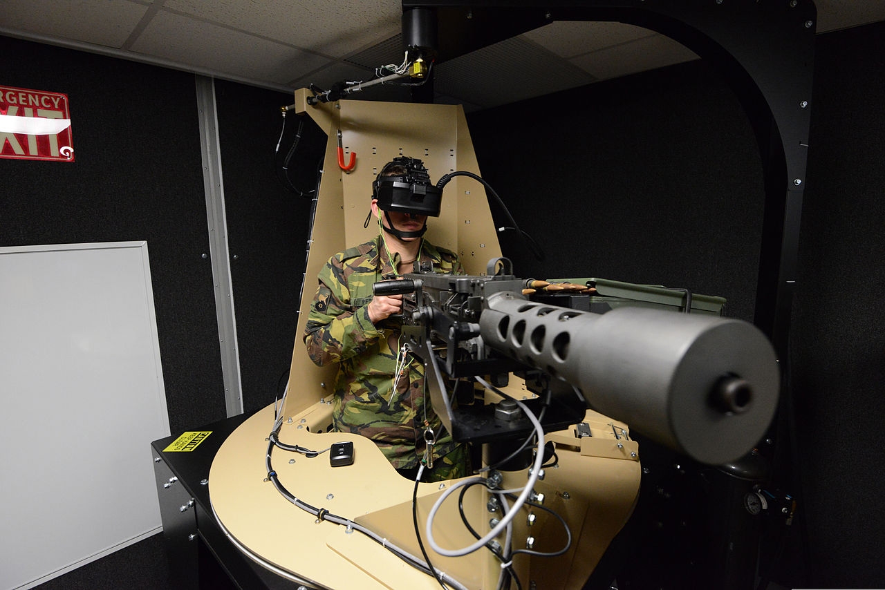 Colour photograph of a man wearing green camouflage clothing with a headset over his eyes and holding a device shaped like a large machine gun and covered in wires.