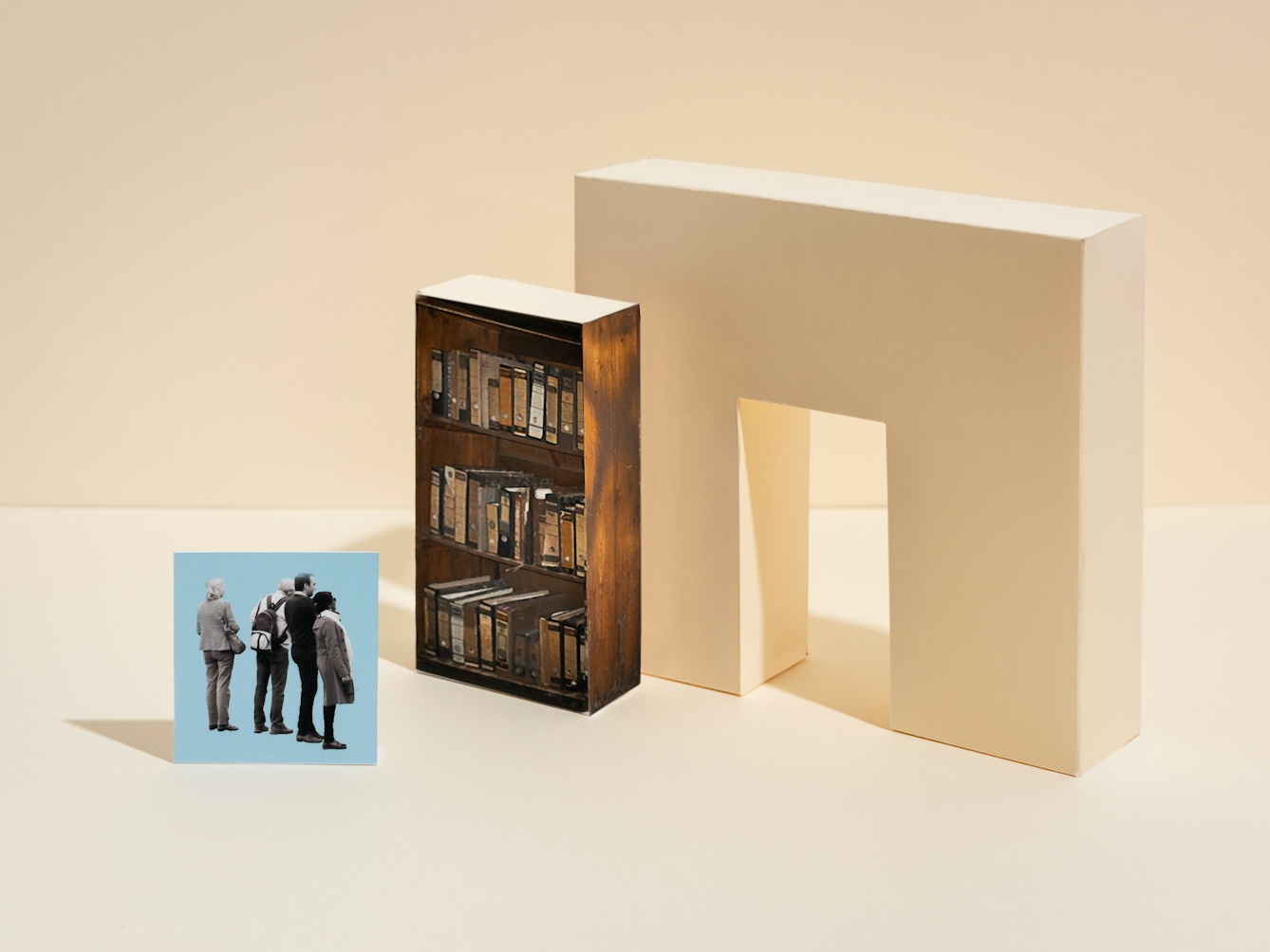 Photograph of a set built scene. The scene is made out of a light cream horizontal surface against a light cream vertical background with a thin horizon line. On the horizontal surface are two three-dimensional shapes. The shape on the right is a light cream wall with a doorway though the centre. To the left of the doorway is another rectangular cuboid which is covered in a printed surface showing 3 book shelves containing archive lever arch folders. To the left of the bookshelf is a rectangular flat blue card standing upright, on which is the black and white image of a group of people from behind looking towards the book shelf.
