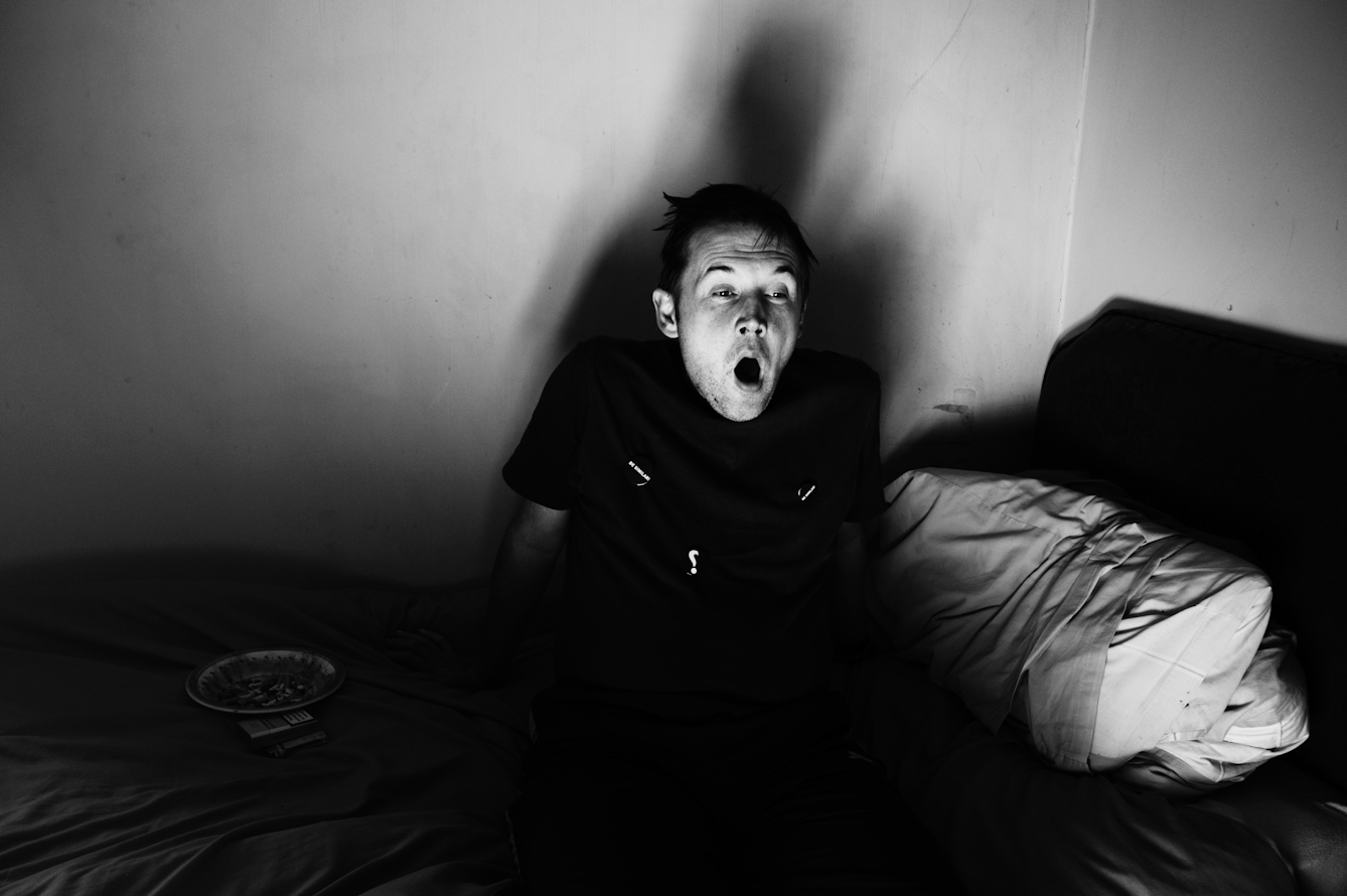 Black and white photograph showing a man sitting sideways on a bed, yawning. The lighting in the room is from a low angle casting his shadow on the wall. Beside him on the bed is a plate of food.