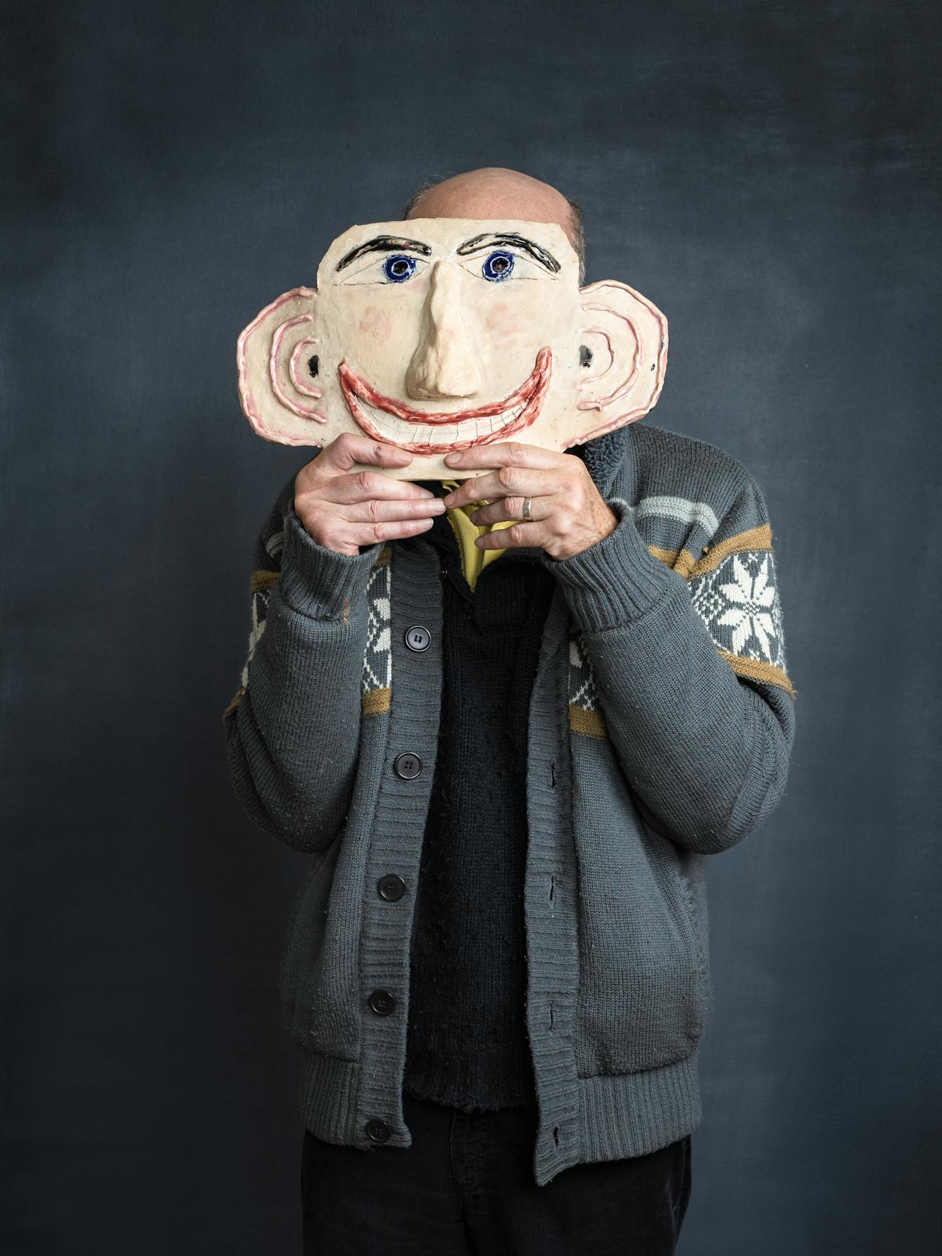 Photograph of stroke survivor and artist Chris Miller in an artist studio. A grey textured background frames the artist as he holds a ceramic of a head, which acts as a cartoon likeness of his own face and which obscures his own face. 