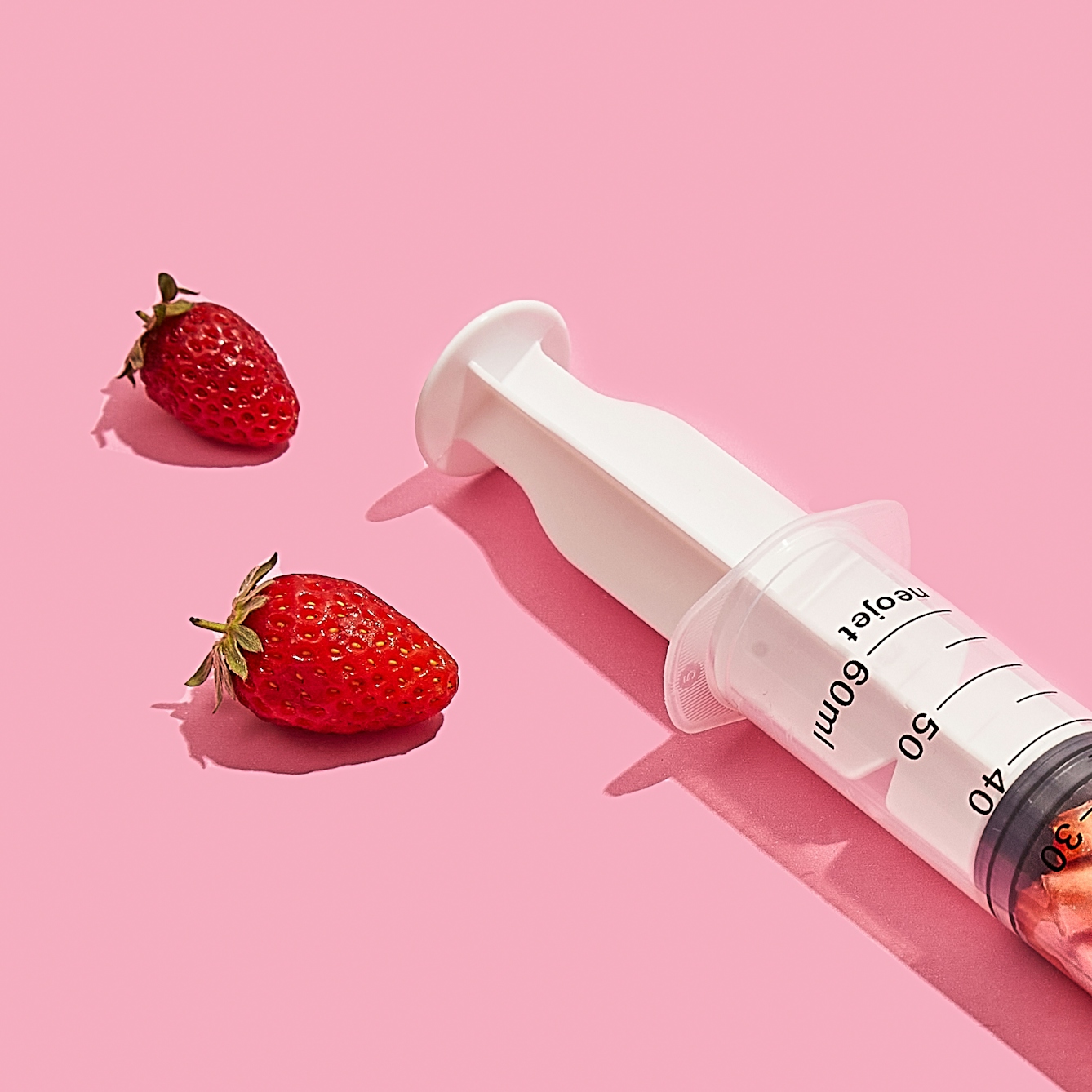 Photograph of a medical syringe lying on its side on a bright pink background. At the plunger end of the syringe are 2 strawberries lying on their side on the background.