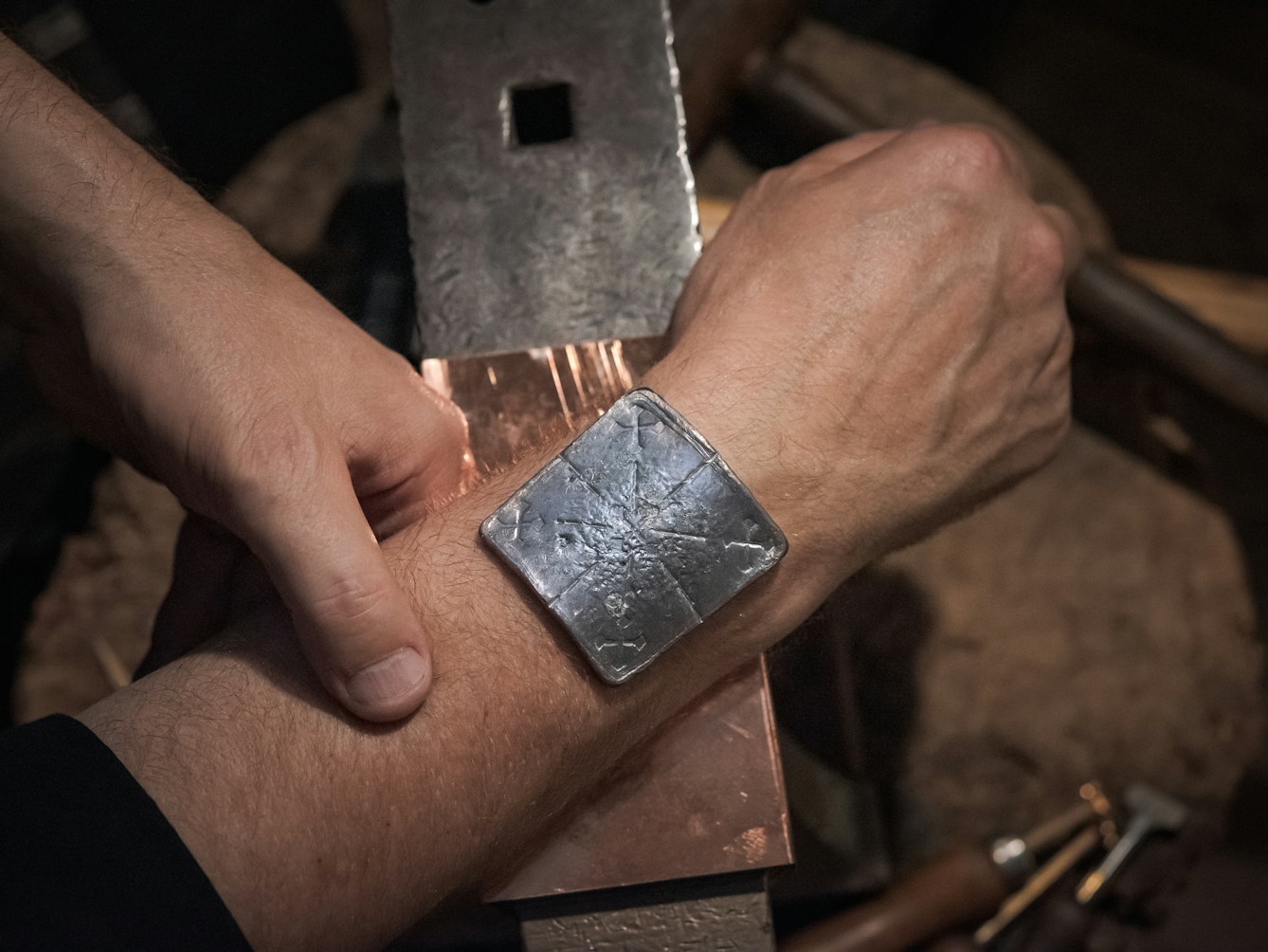 Photograph of the lead healing plate placed on the wrist of a man.  The wrist is resting on an anvil. Inscribed on the lead plate are five crosses, representing the five wounds of Christ. 
