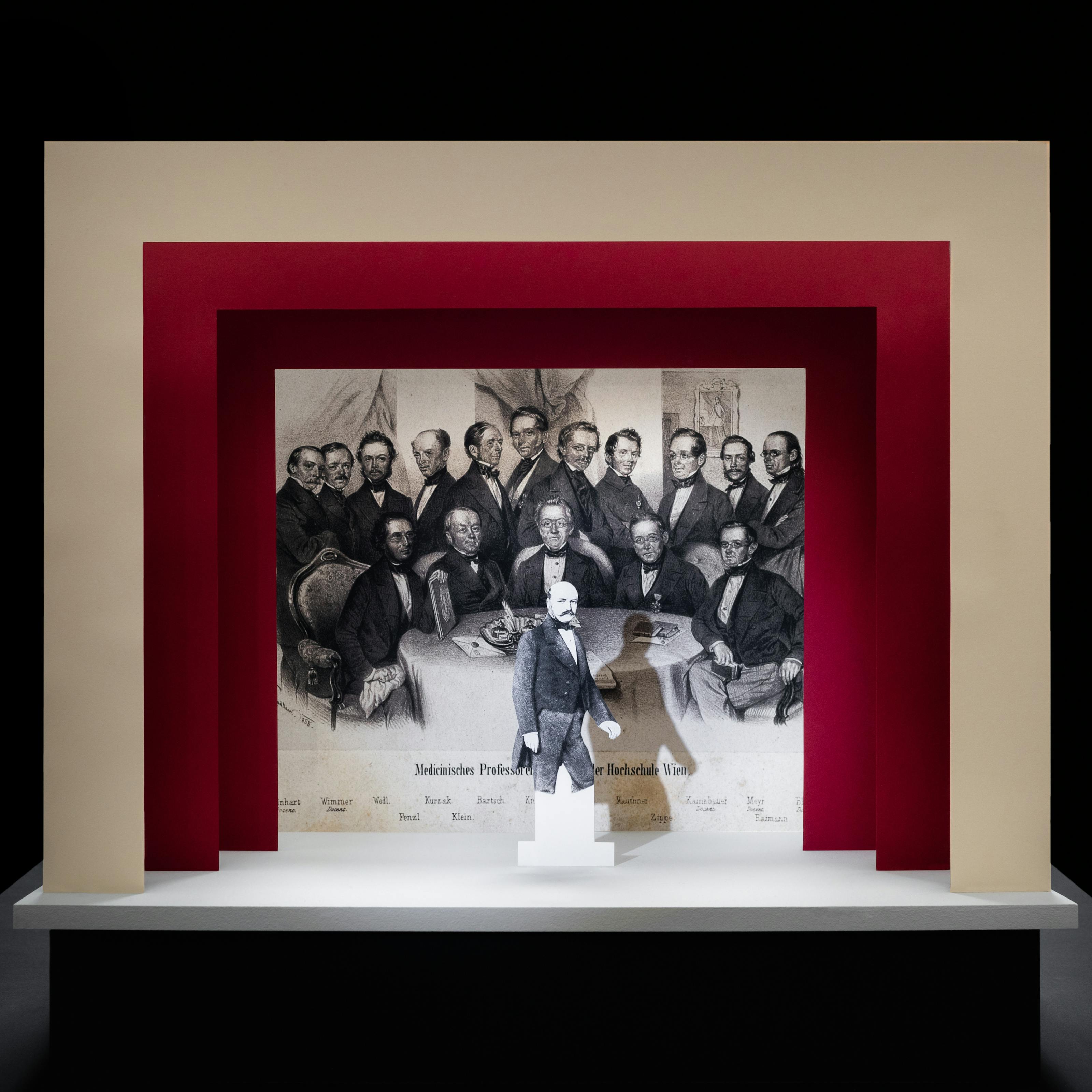 Photograph of a simple theatre stage set, made out of card. The background surrounding the stage is black. The stage floor is white and the framing of the stage is made out of 3 square edged arches, each one smaller than the other, receding backwards. The first arch is cream coloured and the other two are a red. On the stage is a small cut out illustration of a man with a moustache from the early 20th century. Behind him forming the backdrop is a black and white drawing of a group of suited men gathered around a table looking towards the viewer. Under these men are a list of their names.