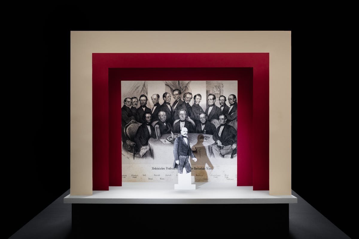 Photograph of a simple theatre stage set, made out of card. The background surrounding the stage is black. The stage floor is white and the framing of the stage is made out of 3 square edged arches, each one smaller than the other, receding backwards. The first arch is cream coloured and the other two are a red. On the stage is a small cut out illustration of a man with a moustache from the early 20th century. Behind him forming the backdrop is a black and white drawing of a group of suited men gathered around a table looking towards the viewer. Under these men are a list of their names.