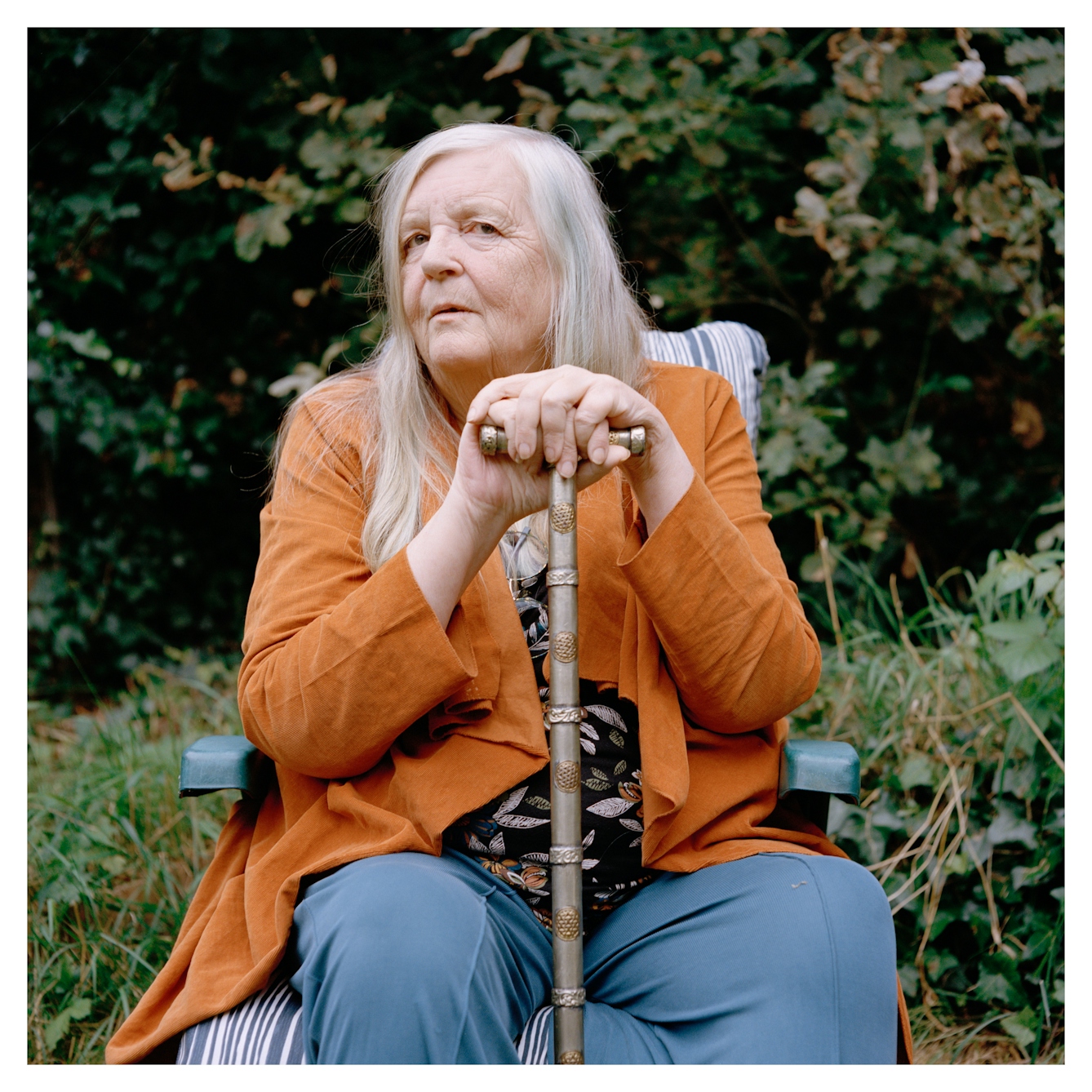 Portrait photograph of Alison, a woman with long grey hair, sitting on a plastic garden chair. Her hands are both resting on a silver walking stick, one hand stacked on top of the other. She is wearing blue trousers, a patterned top and an orange jacket. She has a neutral expression and is looking at the camera. Behind her are are some trees and small plants. 