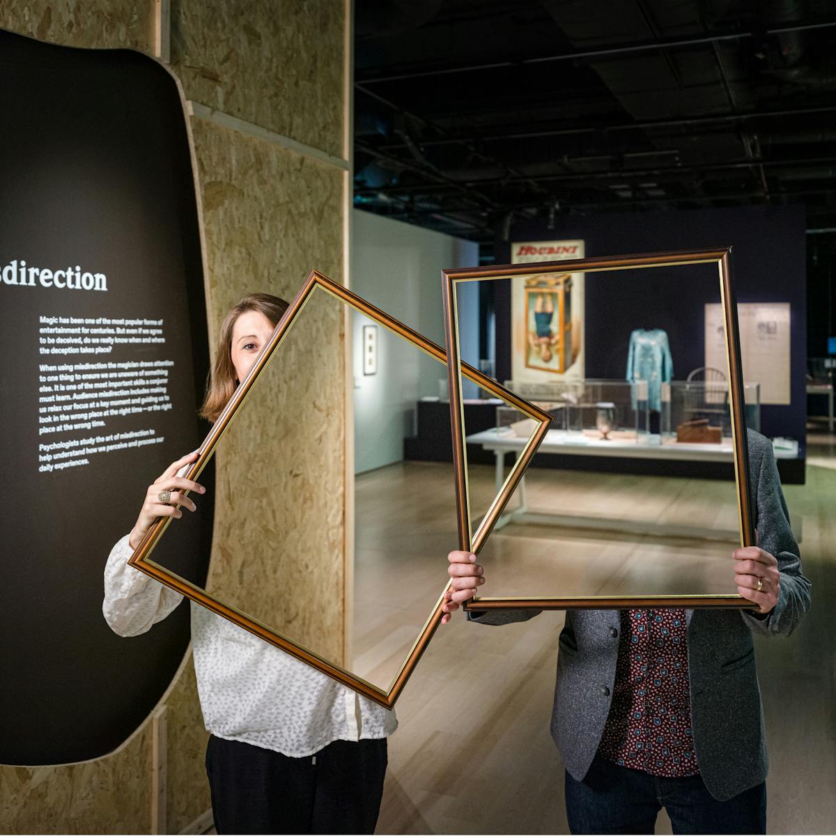 Photograph of a woman and a man in a dark gallery space holding up picture frames in front of their bodies and parts of their faces. Within the picture frame you can see straight through them, as if their bodies and faces have disappeared. In the background you can see a large historical poster and exhibitor display cases. On the left is a text panel titled, "Misdirection".