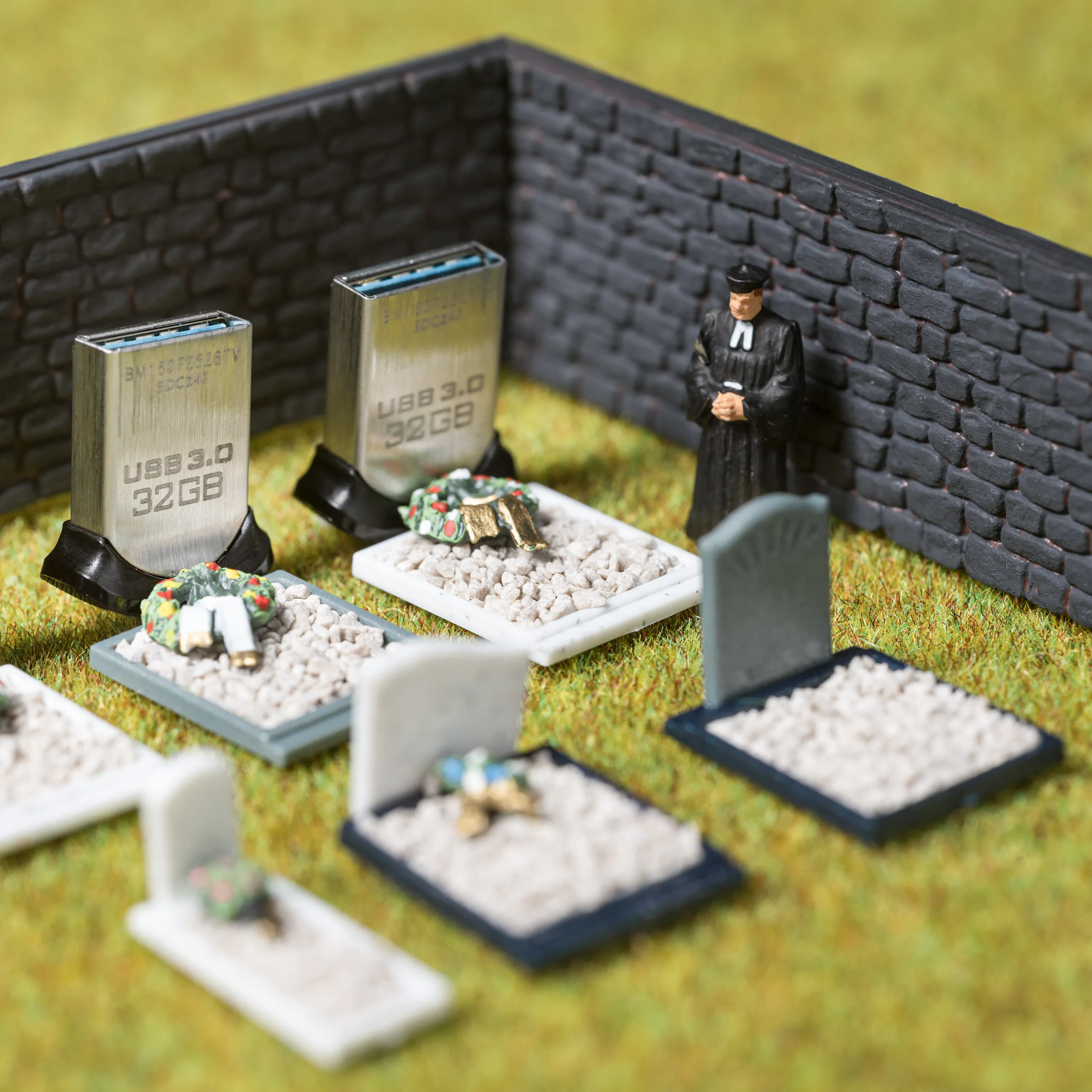 Photograph of model railway scenery depicting a walled cemetery containing seven graves. Models of a man holding flowers and a clergyman strand over the graves. Three of the headstones are made of actual size USB memory sticks standing on their ends. On the sticks is the text, USB 3.0, 32GB.
