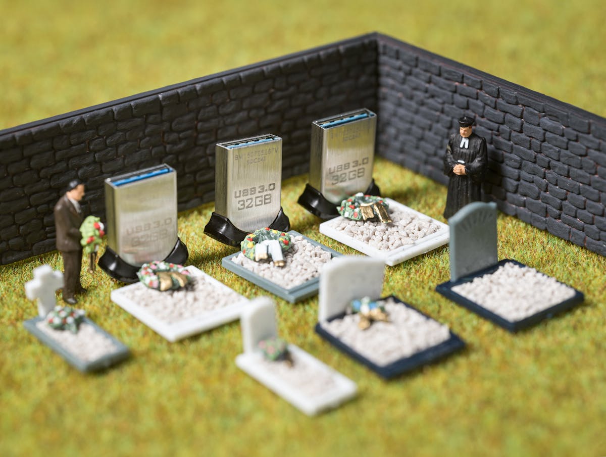 Photograph of model railway scenery depicting a walled cemetery containing seven graves. Models of a man holding flowers and a clergyman strand over the graves. Three of the headstones are made of actual size USB memory sticks standing on their ends. On the sticks is the text, USB 3.0, 32GB.