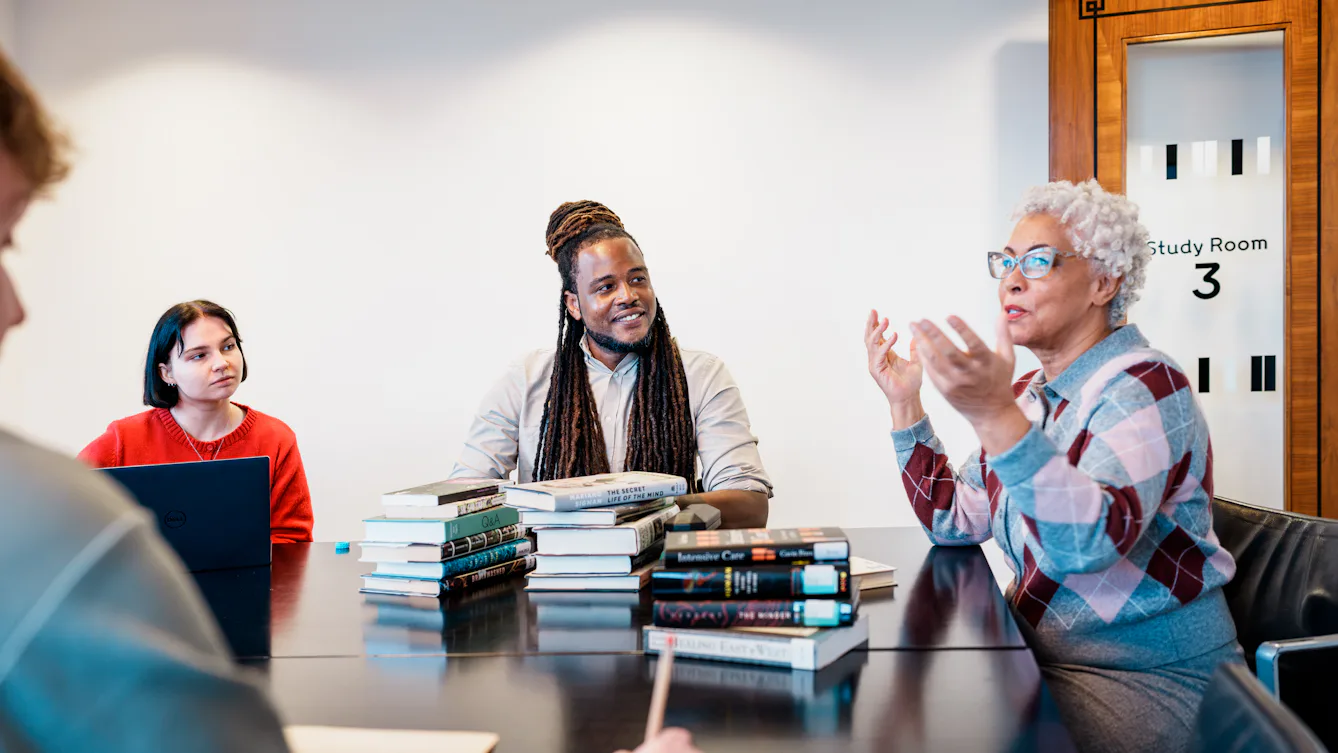 Photograph of a group of 4 library users sat around a desk in discussion. On the table are stacks of books, note pads and a laptop.