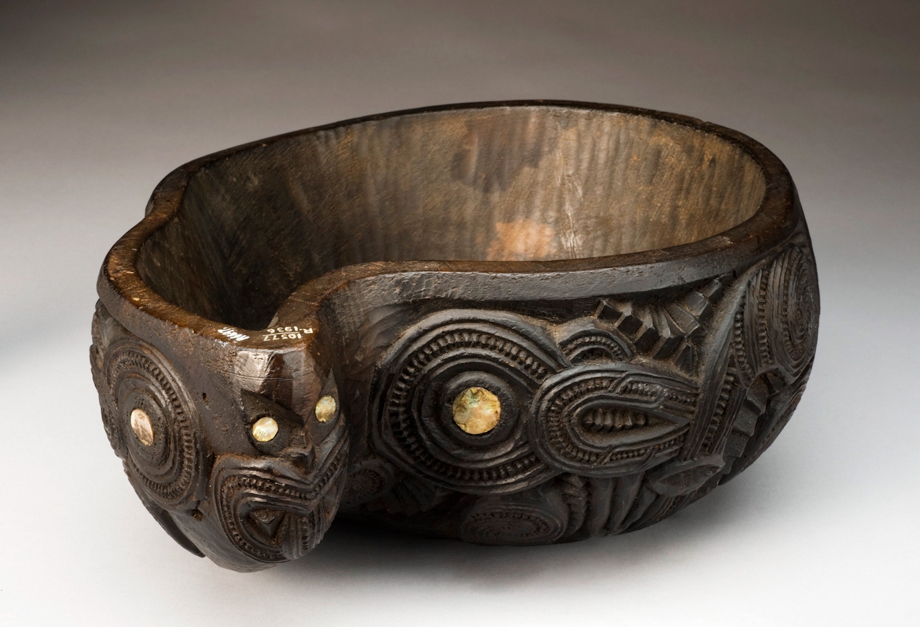 A photograph of a carved wooden bowl inset with abalone shell. The bowl is of New Zealand origin and shows traditional Maori overlapping animals and ancestral figures called Tiki.