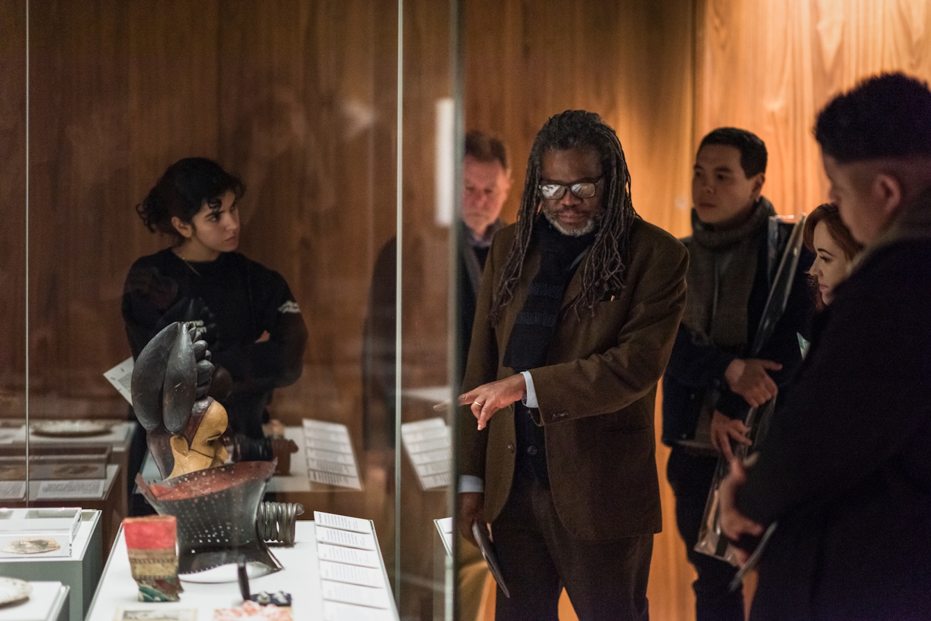 Photograph of a man in a dark gallery space in front of a display case, pointing towards an object inside. He is surrounded by a small group of people who are listening to what he has to say.