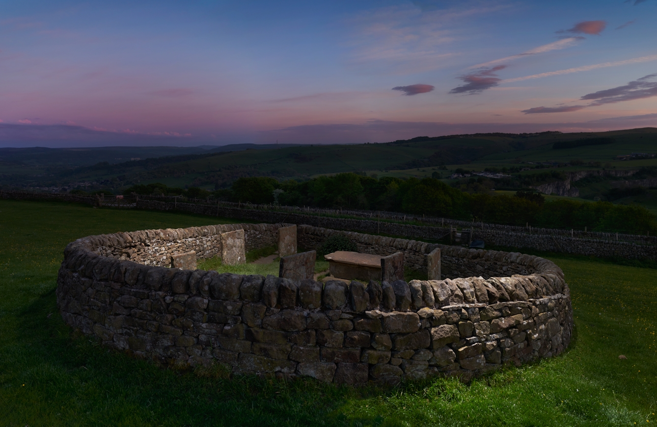 Photograph of rural landscape at dusk, with a circular dry stone walled enclosure, housing several grave stones.