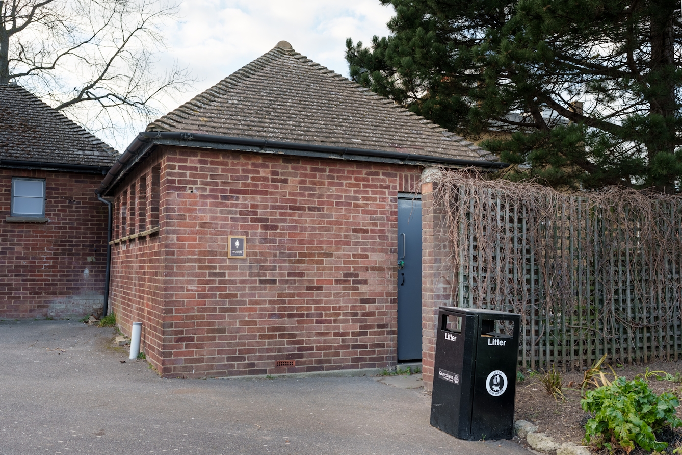 A photograph of a small building that has a shiny metal sign with a figure and the text womens. Next to that is a grey door which is padlocked shut.