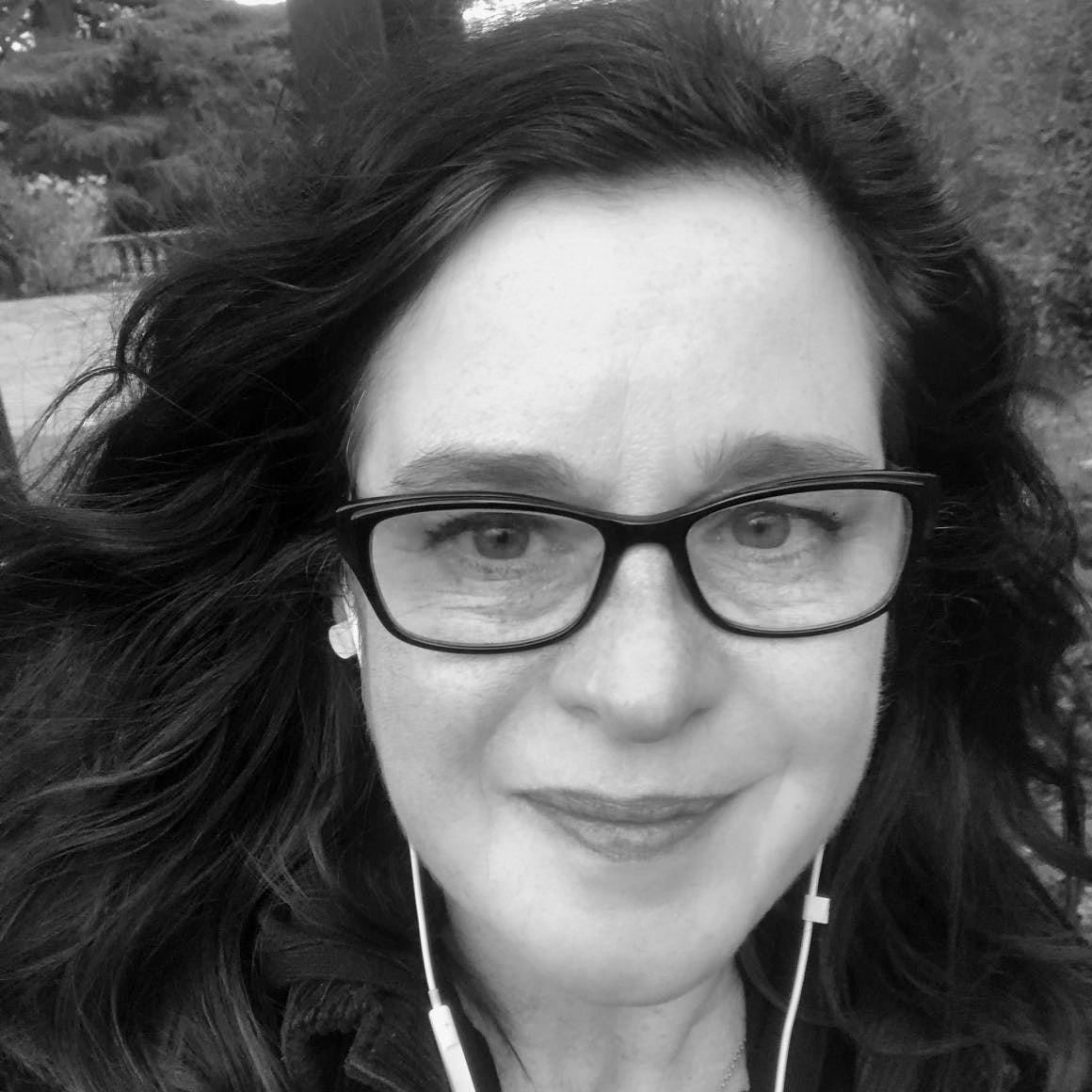 Black and white, head shot of a woman with long dark hair, wearing spectacles and trailing leads from earbuds, against the background of a park.