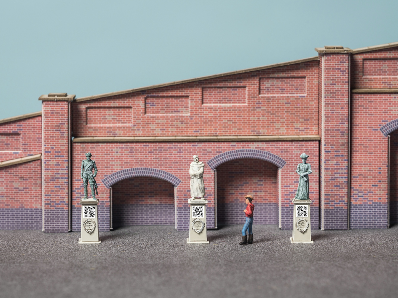Photograph of model railway scenery depicting a tapered brick wall in front of which are three statues on plinths. Under each statue is a carved wreath with their name in the middle and a printed QR code. A model figure stands looking at the statue in the middle.