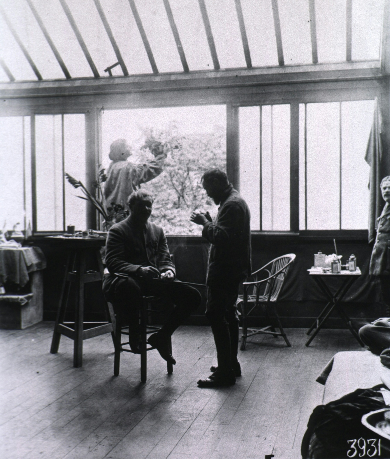 Black and white photograoh of a sunlit stiudio with two standing figures and one seated figure.