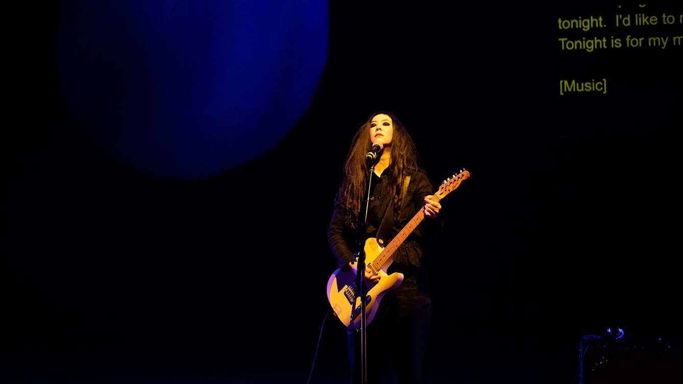 Johanna Hedva performing on stage with a microphone and holding a guitar. 