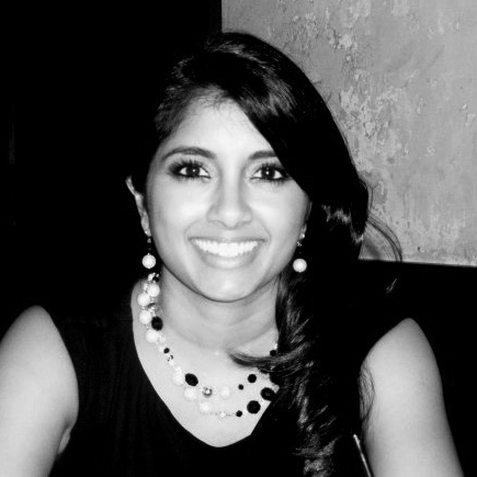 Black and white, head and shoulder photograph of a young woman with long, dark hair. She is wearing a black t-shirt with matching necklace and earrings made from large beads. The background is black with a grey square in one corner. 