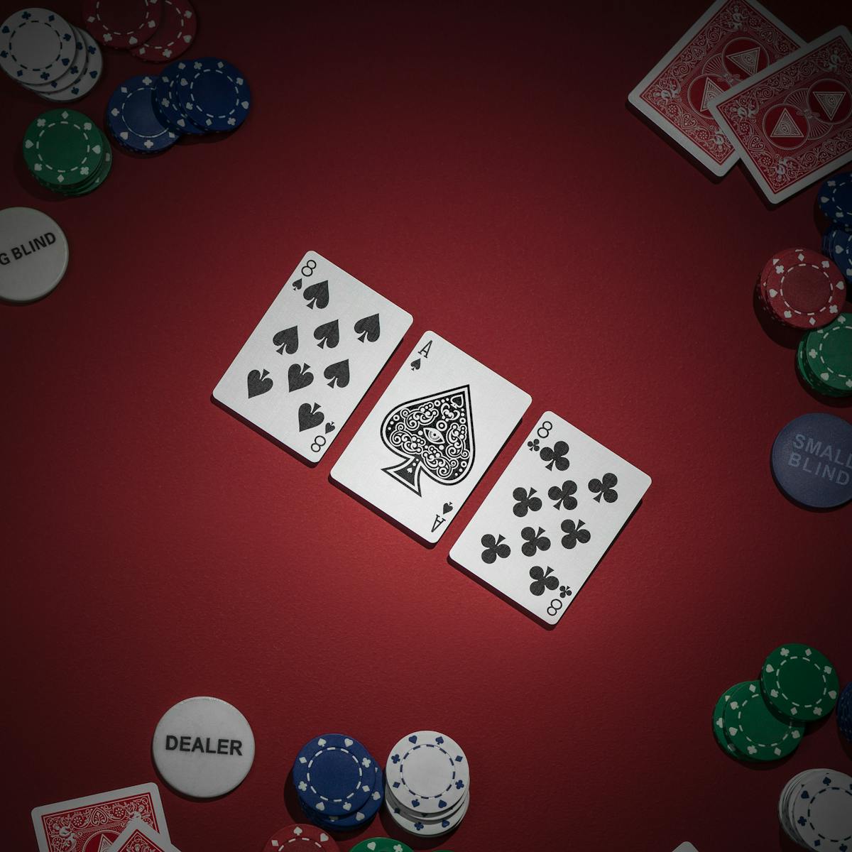 Photograph of a game of texas hold'em poker. Three cards reading eight of spades, ace of spades and eight of clubs have been dealt face up. To the side are four other hands of two cards which are hidden.  A pack of cards waiting to be dealt is visible in the bottom left of the frame.  Chips are present for each player, along with the dealer, big blind and small blind tokens.  The lighting is moody and picks out the upturned cards. The ace has been decorated with the eye of the illuminati.  The backs of the players cards also show the eye of the illuminati in a pyramid.