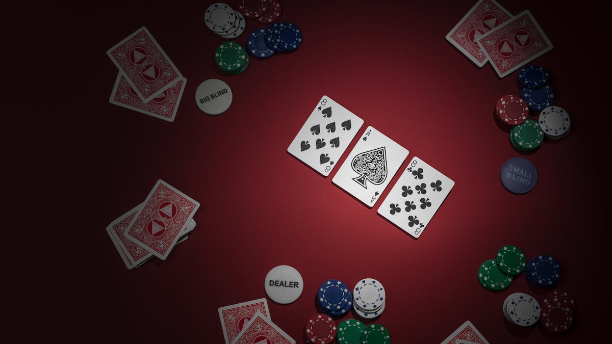 Photograph of a game of texas hold'em poker. Three cards reading eight of spades, ace of spades and eight of clubs have been dealt face up. To the side are four other hands of two cards which are hidden.  A pack of cards waiting to be dealt is visible in the bottom left of the frame.  Chips are present for each player, along with the dealer, big blind and small blind tokens.  The lighting is moody and picks out the upturned cards. The ace has been decorated with the eye of the illuminati.  The backs of the players cards also show the eye of the illuminati in a pyramid.