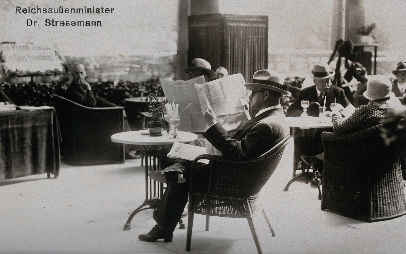 Photograph of  a man sitting at a cafe-style table among others, wearing a pale hat and reading a newspaper.