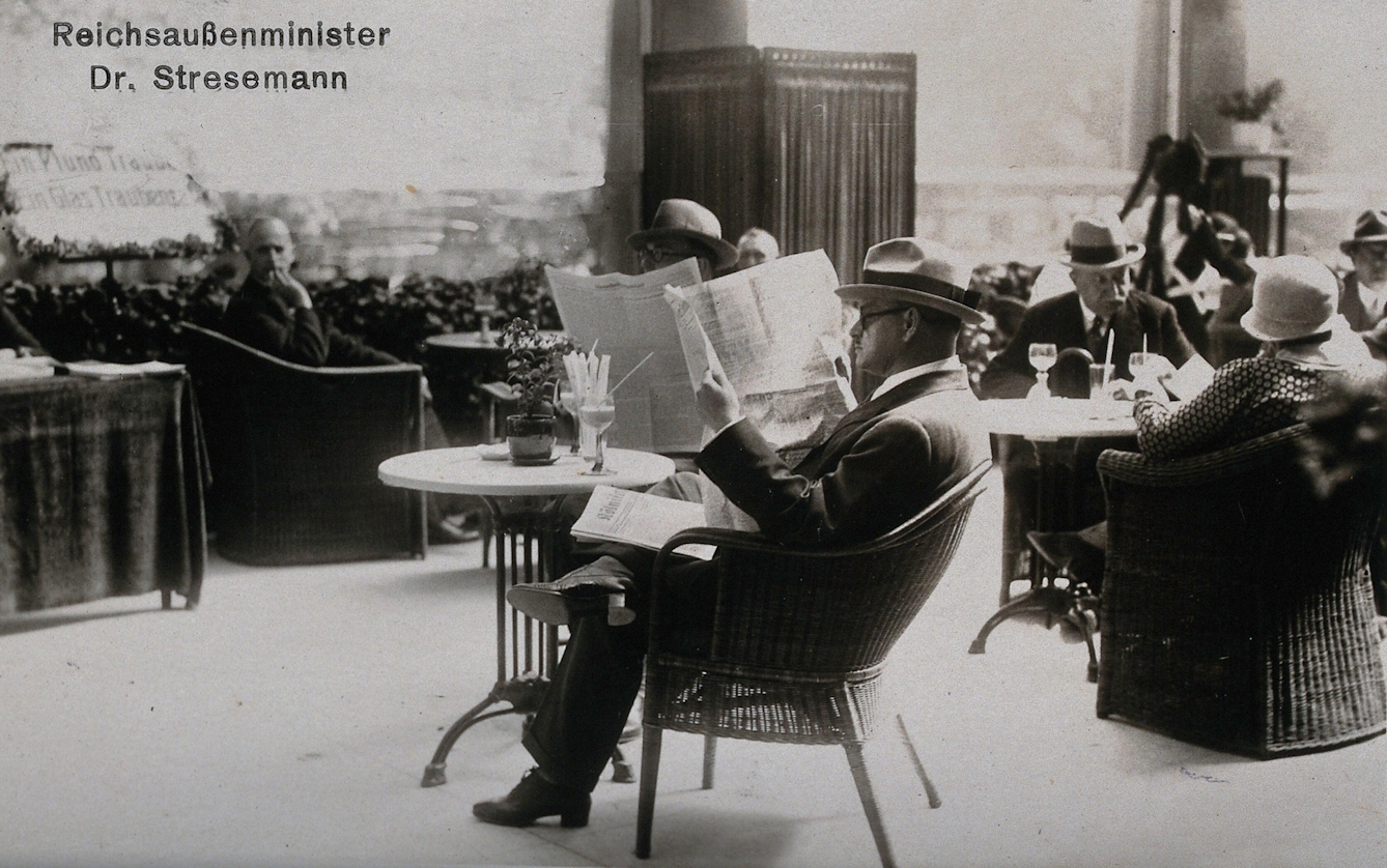 Photograph of a man sitting at a cafe-style table among others, wearing a pale hat and reading a newspaper.