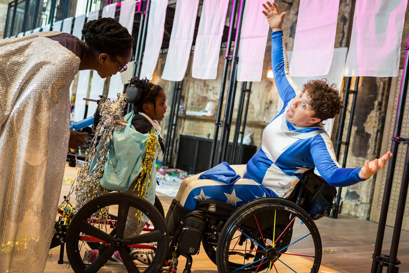 Photograph at an event showing a woman in a wheelchair wearing a blue and white superhero style outfit, with her arms outstretched and her mouth wide open. A mother and daughter are stood in front of her, watching.