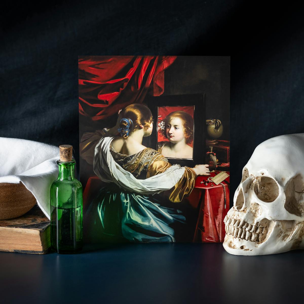 A still life photograph showing a reproduction of a painting depicting a renaissance women sat her dressing table, looking at her reflection in a mirror. She is draped in fine clothes depicting her wealth, with bold red curtains in the back ground. Surrounding the painting are objects including a skull, a dark green apothecary bottle, a book, a bowl and a candle. The scene is presented against a dark cloth background.