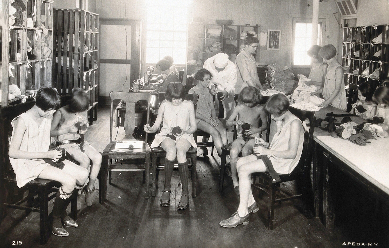 Black and white photograph of a workroom in Mont Alto Sanatorium for Tuberculouis showing adolescent girls sitting darning socks, using serwing machines and standing sorting clothes in the laundry. 1940s.
