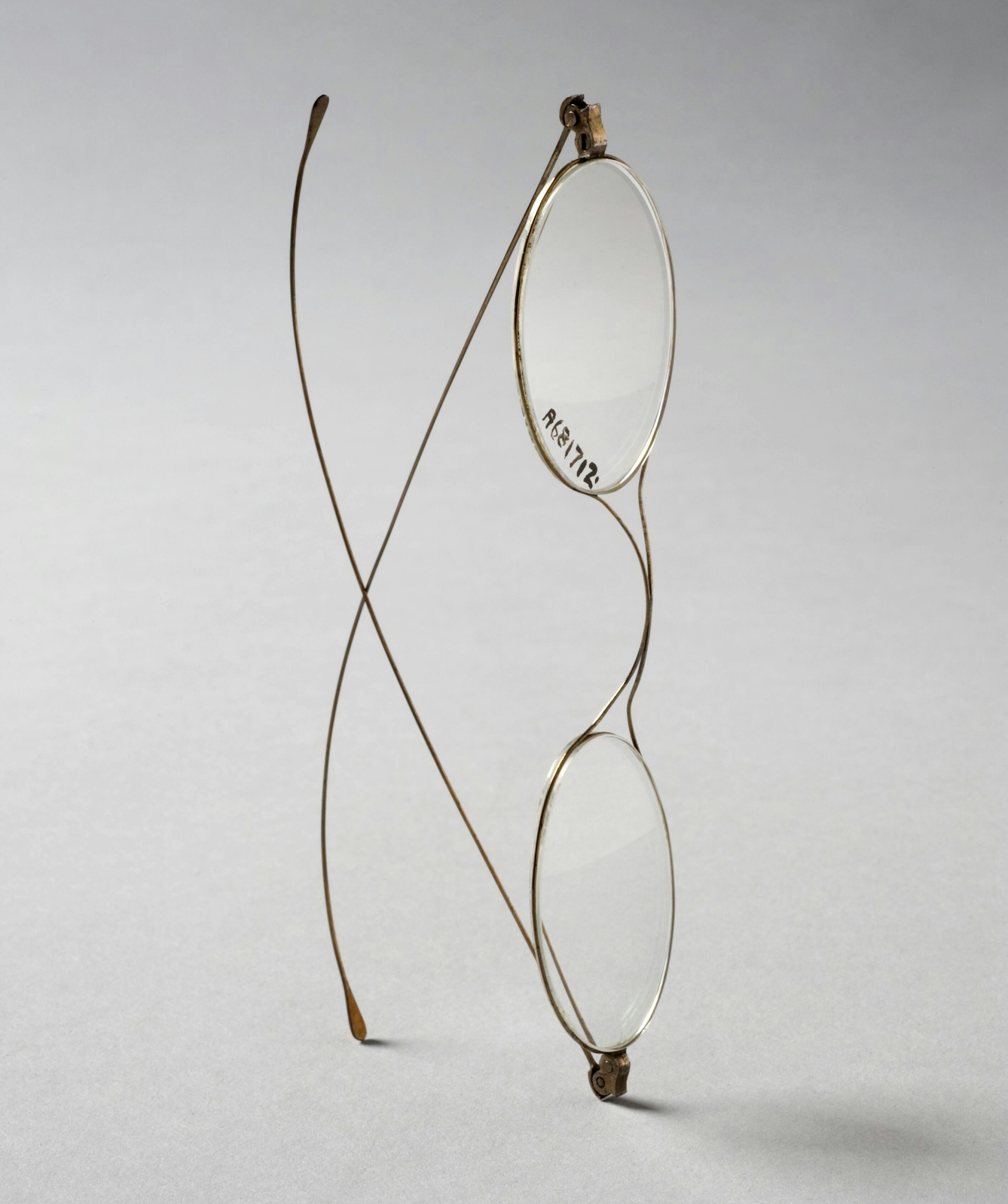 A pair of very thin wire frame spectacles from the 19th century, shown balanced vertically on one arm and one end of the frame and the two arms crossing