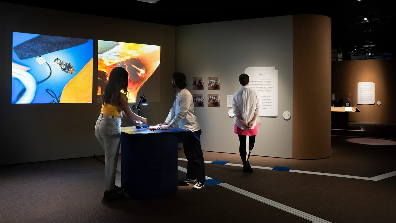 Photograph of three people exploring an exhibition. Two people are stood facing each other over a tabletop. On the table are a selection of object which they are arranging under a camera. In front of them on the gallery wall there are two square projections. On the left is the camera image of the objects they have arranged, a watch, cables against a blue and orange background. On the right is a computer-generated interpretation of their arrangement, abstract white, orange and red shapes.