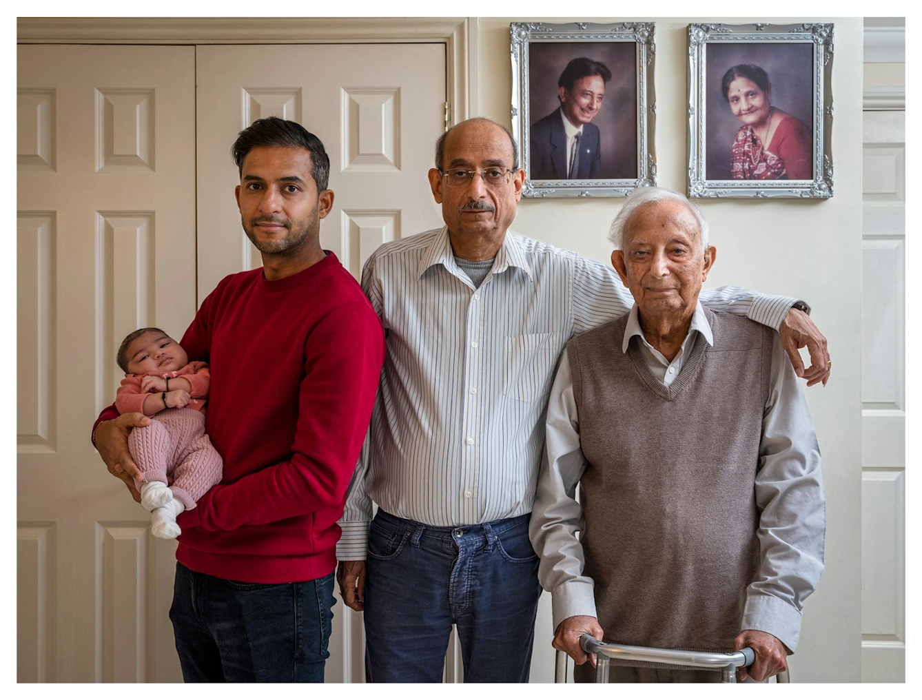 Family group photograph showing four generations of the same family. The great-grandfather stands on the right holding a walking frame, by his side is the grandfather whose arm rests on the great-grandfather's shoulder. Next to him is the grandson who cradles the great-granddaughter who is a baby in his arms. They are stood in front of a white wall on which two studio portraits are hung in silver frames, showing the great-grandfather and his wife many years before. They all look straight into the camera.